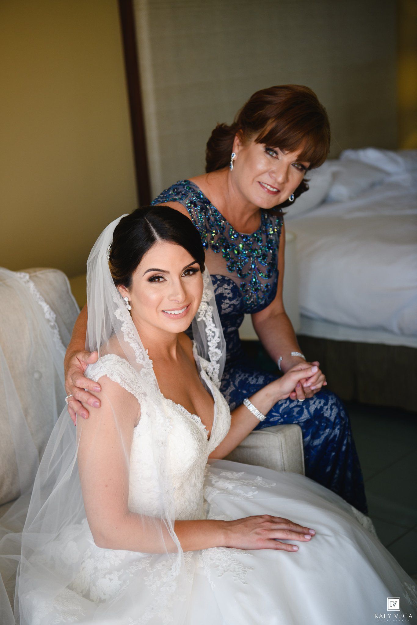 Bride in white lace gown and veil sits with older woman in blue beaded dress during wedding preparations.