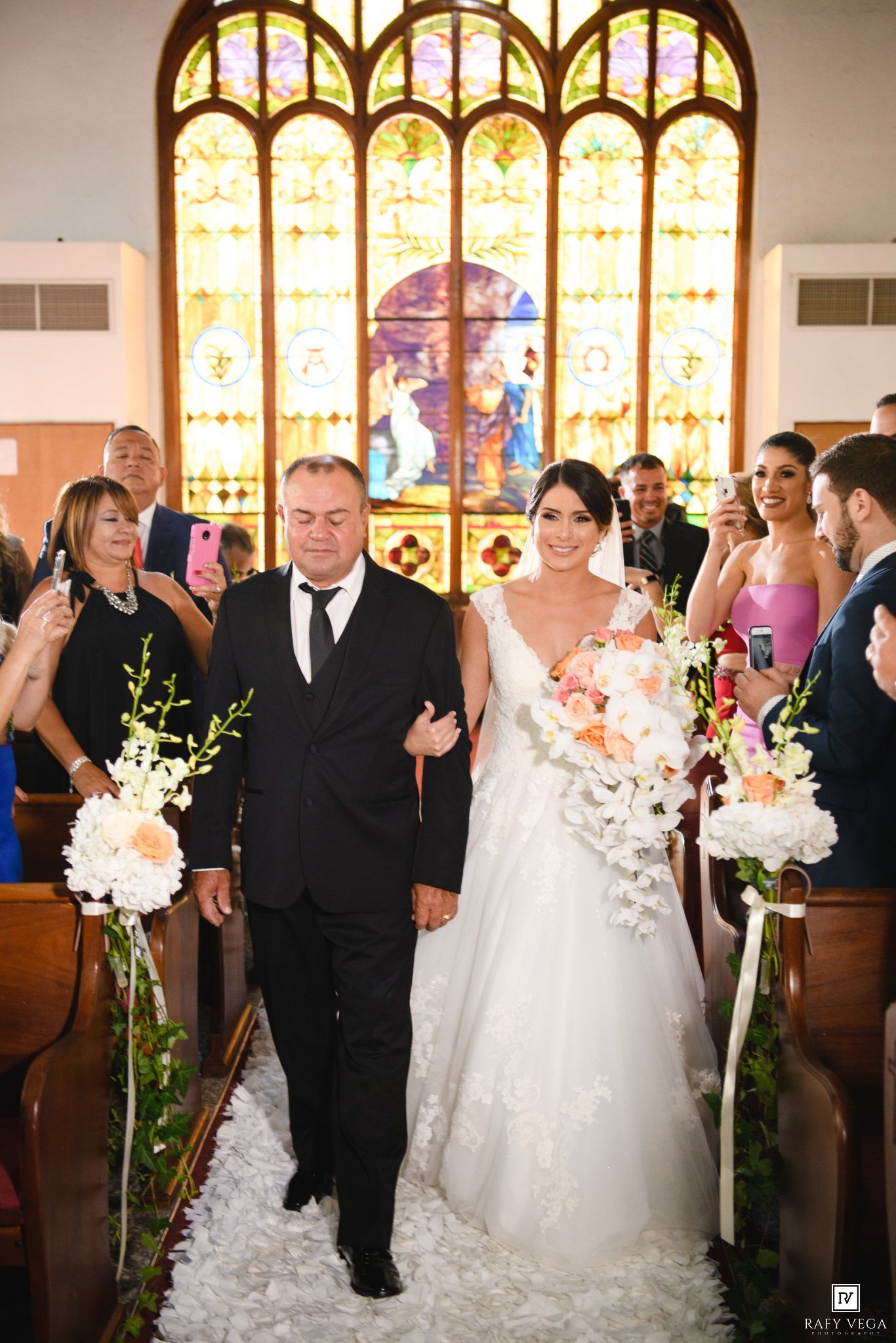 Bride walks down church aisle with escort past floral arrangements, stained glass window backdrop, wedding ceremony.