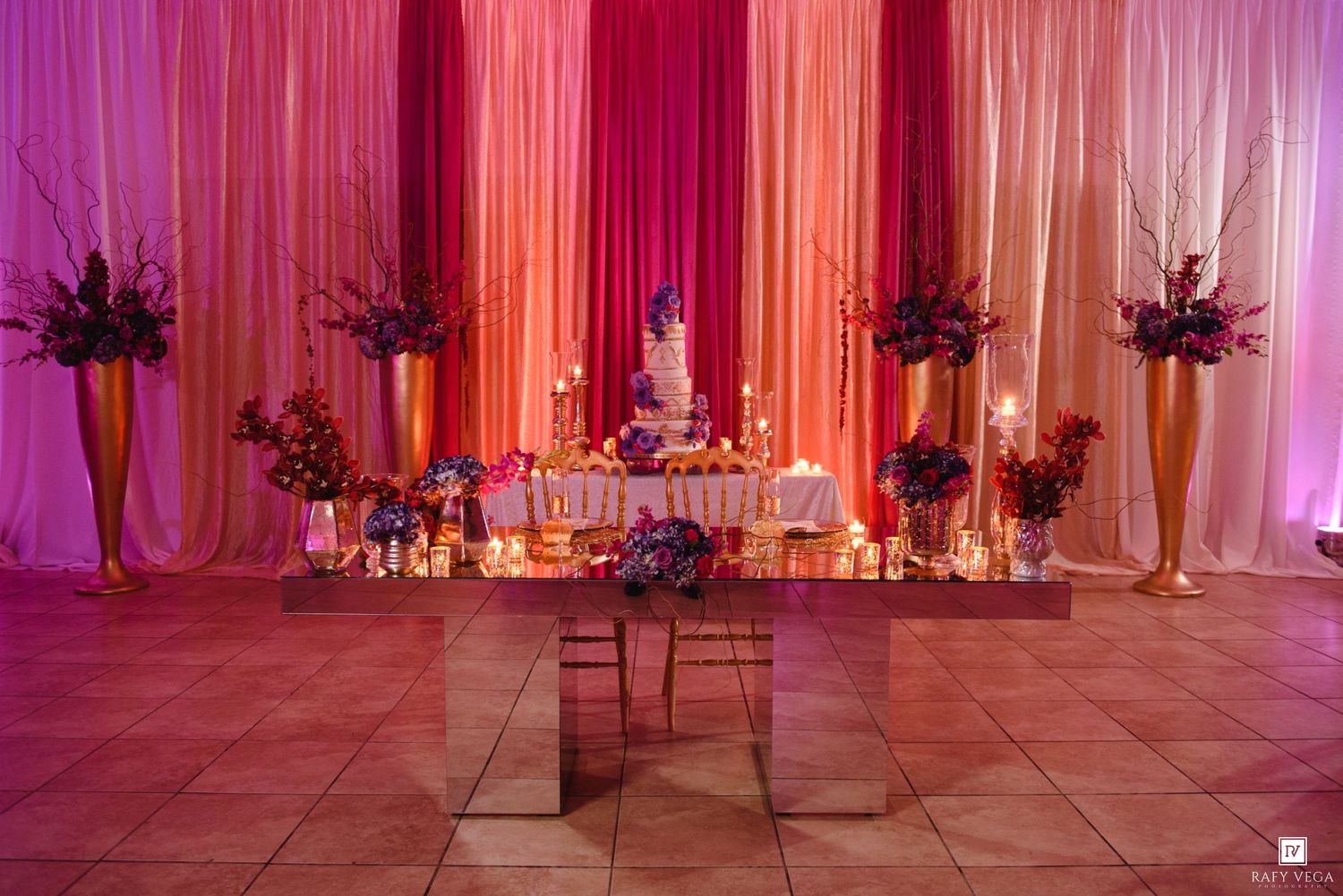 Elegant wedding reception dessert table with pink and orange draping, floral arrangements, candles, and a tiered cake.