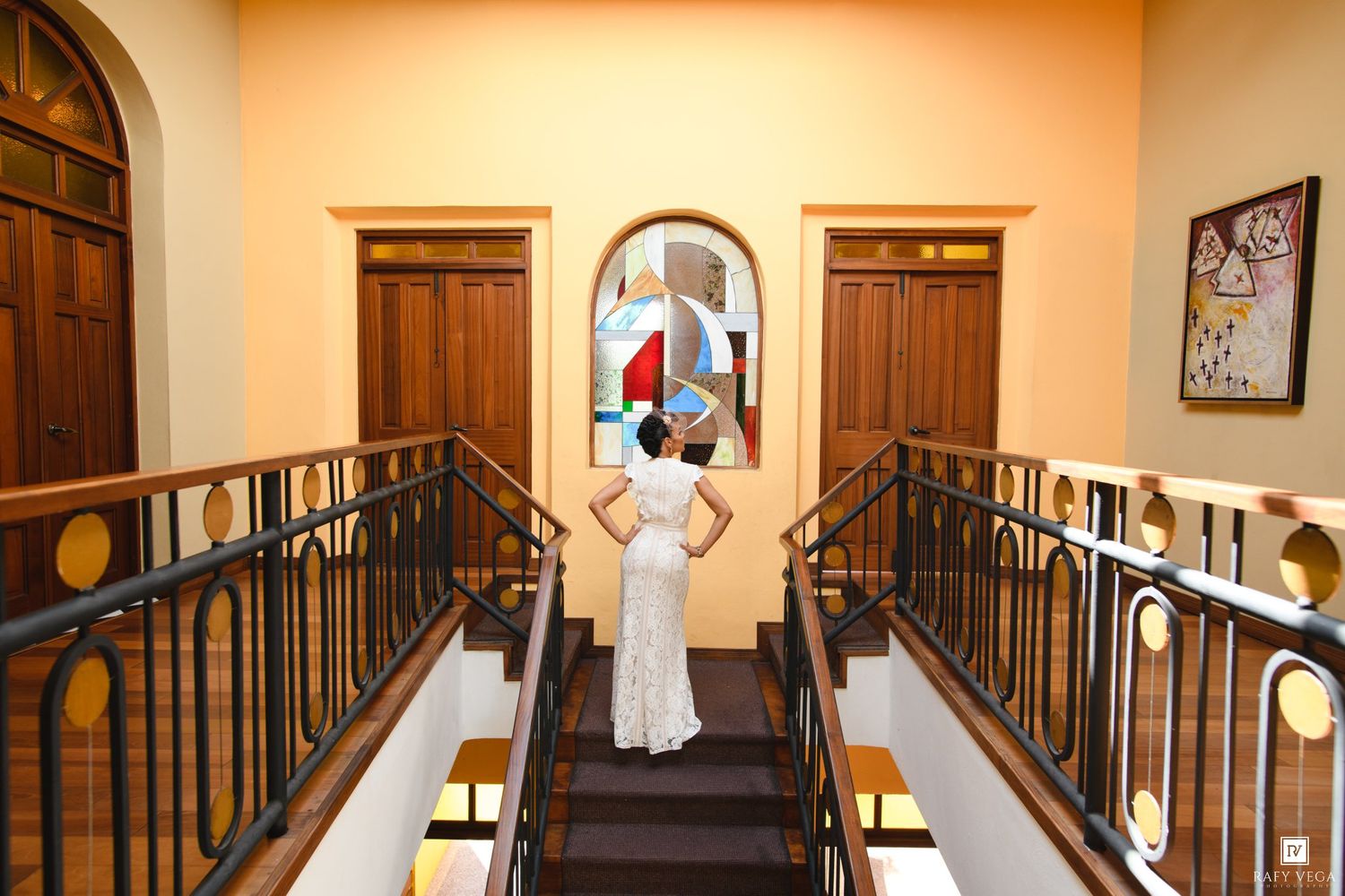 Bride in white gown stands at top of elegant staircase with stained glass window in a classic wedding venue.