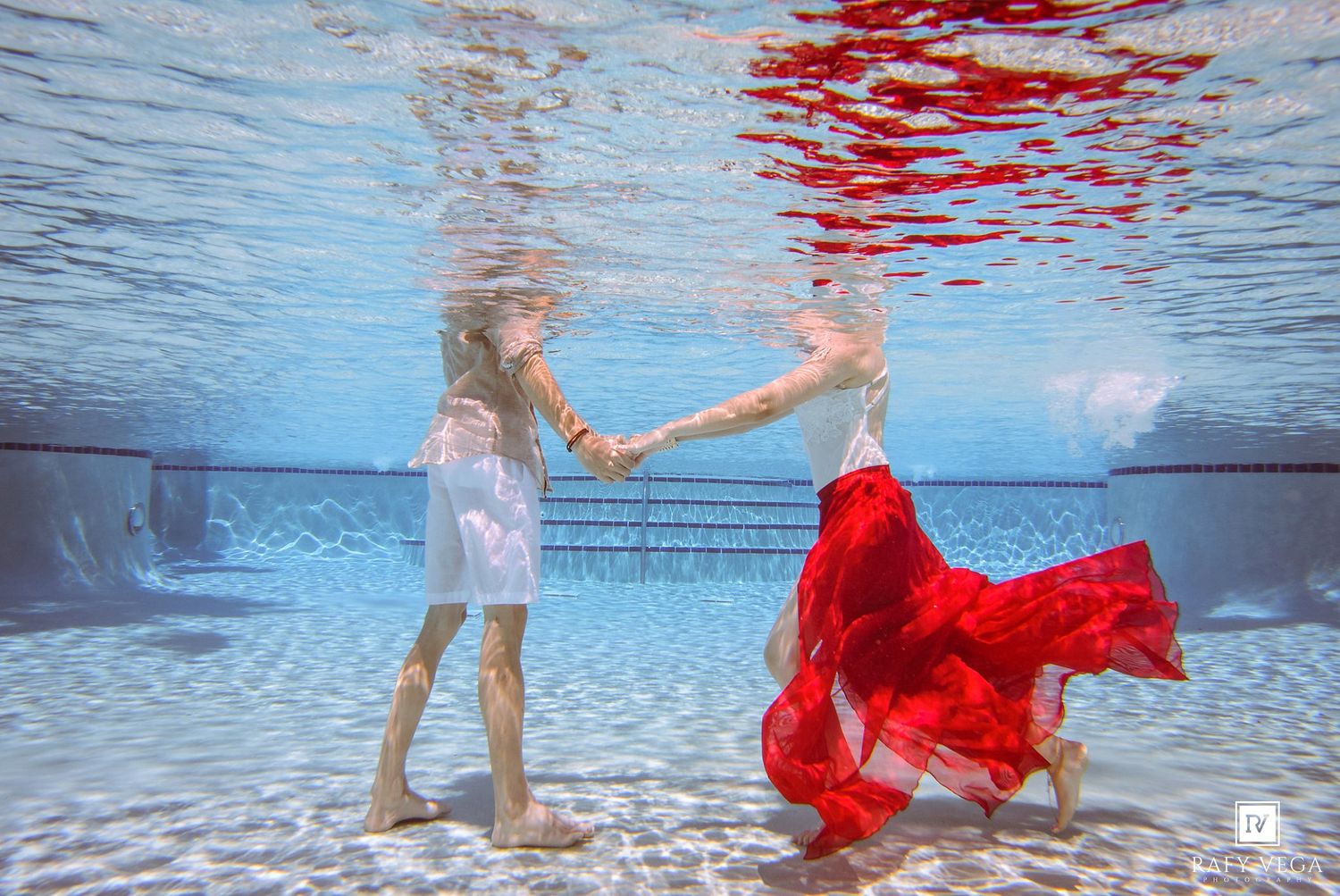 Two girls dancing underwater in a pool, one wearing a white dress and the other a flowing red gown.