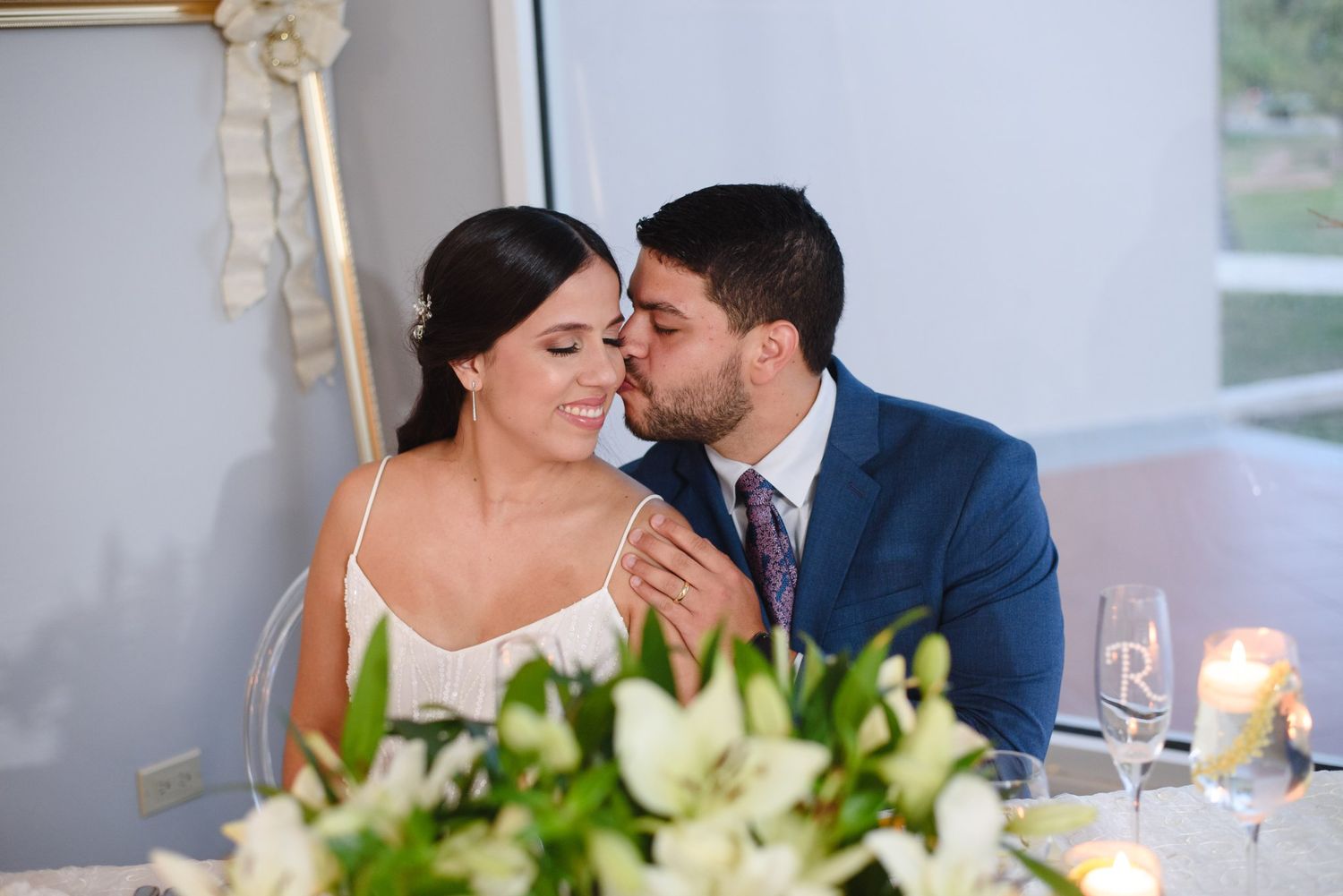 Groom in blue suit kisses bride on cheek at wedding reception table with white flowers and candles.