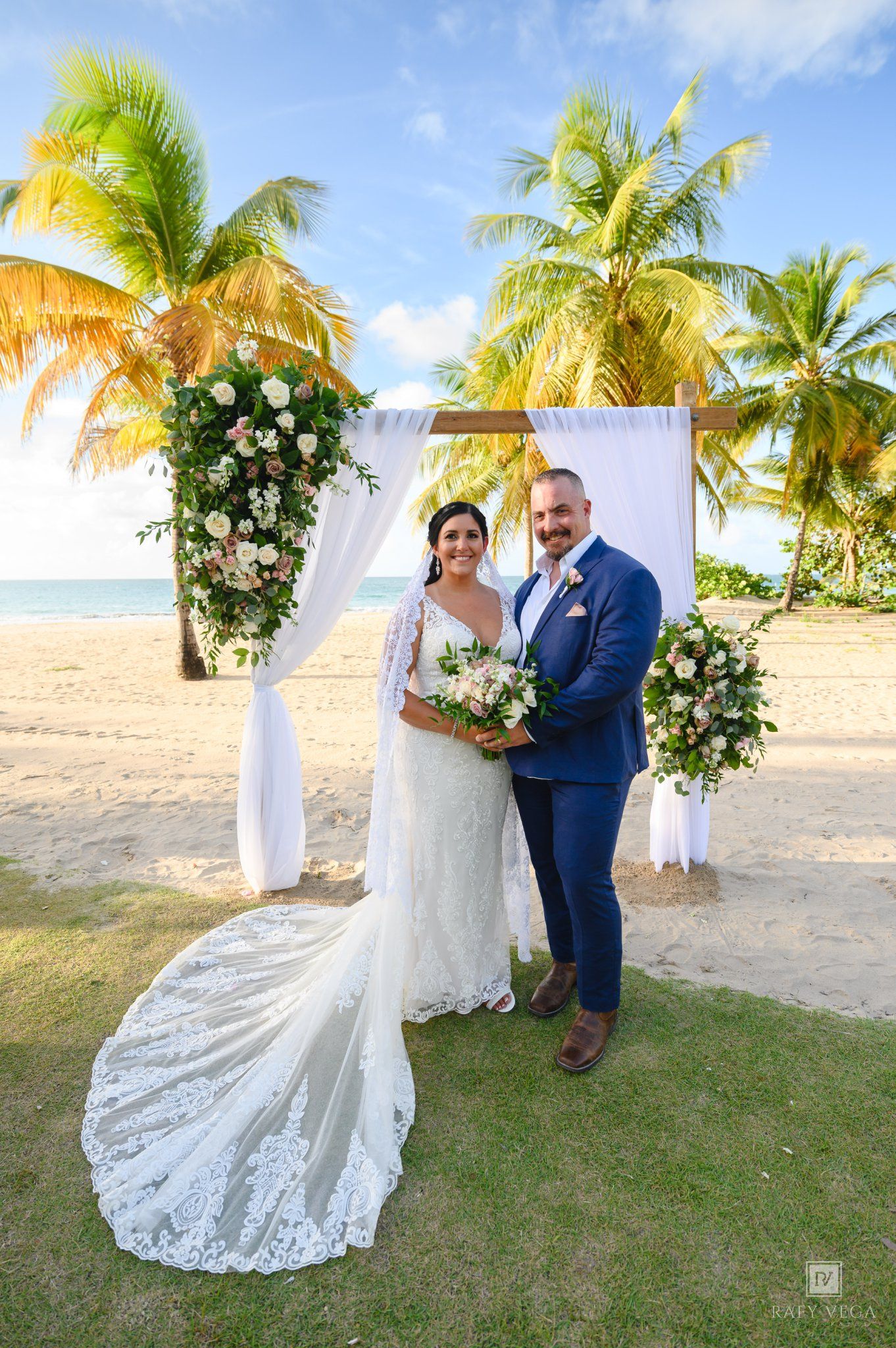 Couple poses at tropical beach wedding ceremony under floral arch with palm trees and ocean backdrop.