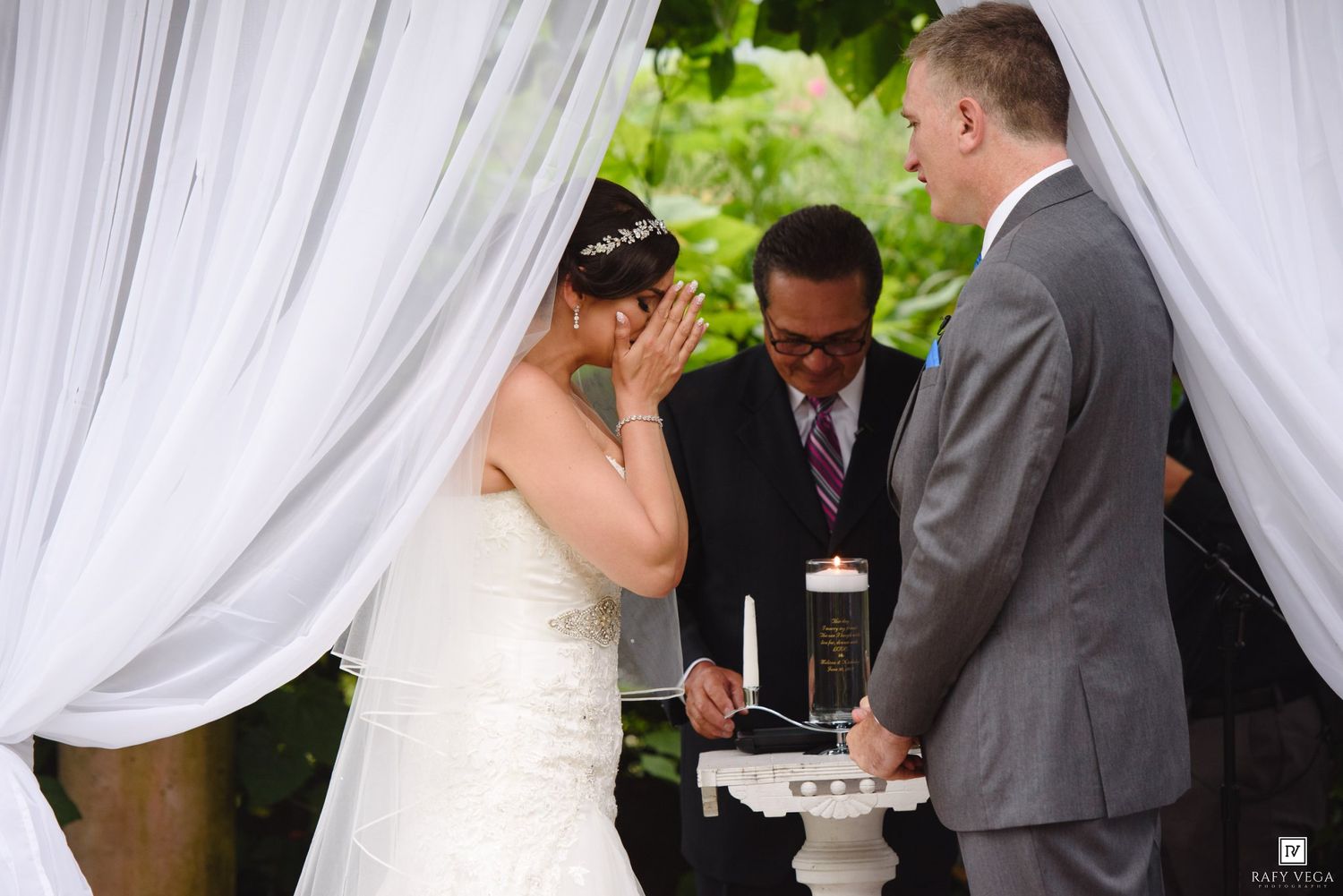 Emotional bride wiping tears during outdoor wedding ceremony with officiant and groom beside white draped canopy.