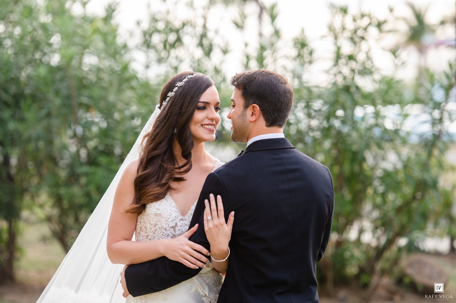 Bride and groom share romantic moment outdoors, bride in lace gown with veil, groom in dark suit.