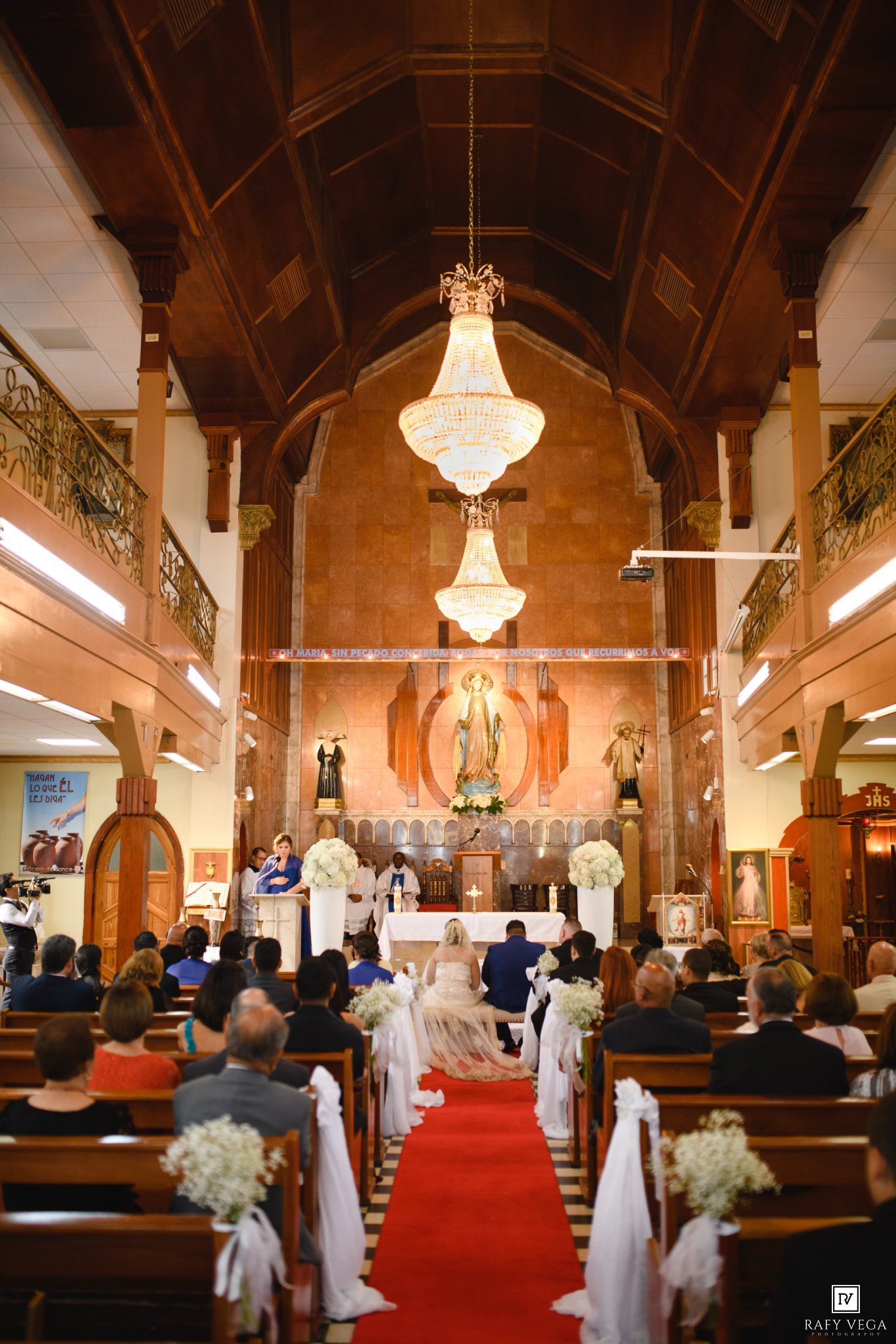 Elegant church wedding ceremony with red carpet aisle, crystal chandeliers, and guests seated in wooden pews.