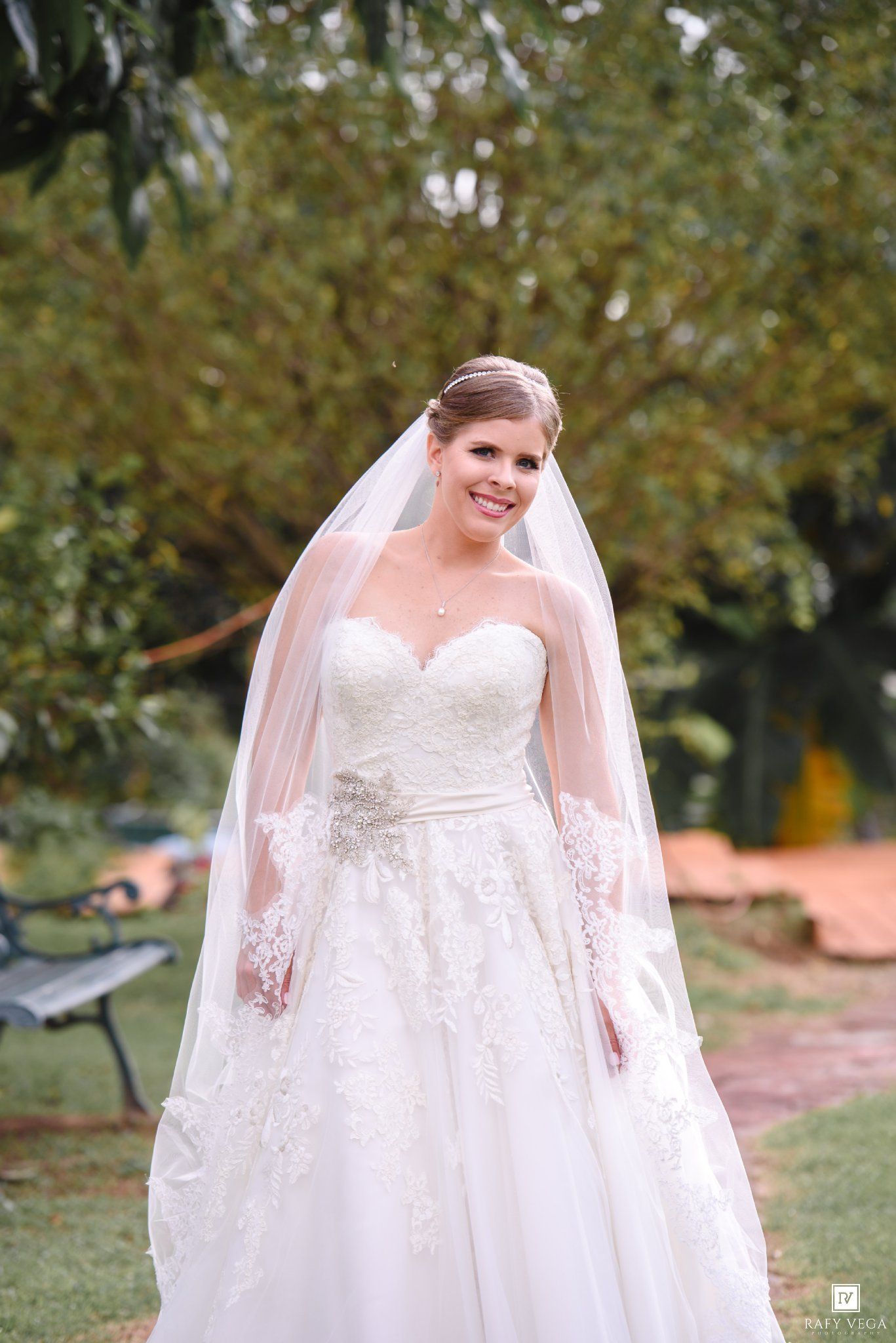 Bride in strapless lace ball gown with veil and crystal belt, smiling outdoors among green trees.