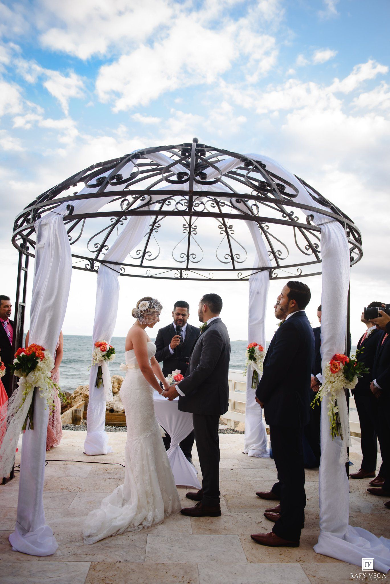 Couple exchanges vows under ornate iron gazebo by ocean at destination beach wedding.