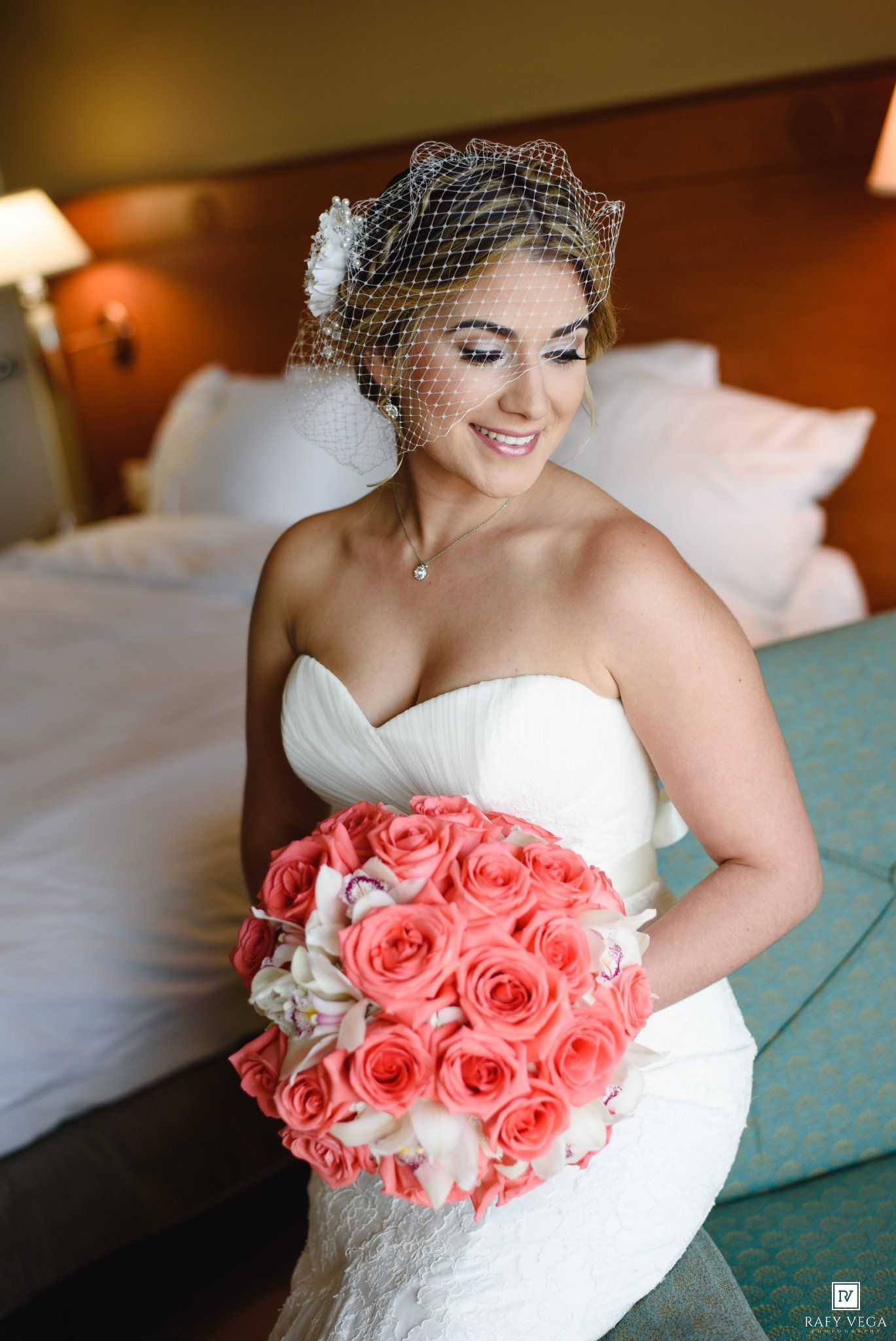 Smiling bride in strapless white dress holding coral rose bouquet, wearing birdcage veil in hotel room.