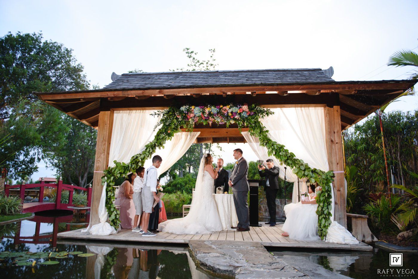 Elegant outdoor wedding ceremony under a floral gazebo by a pond, bride and groom exchanging vows with guests.