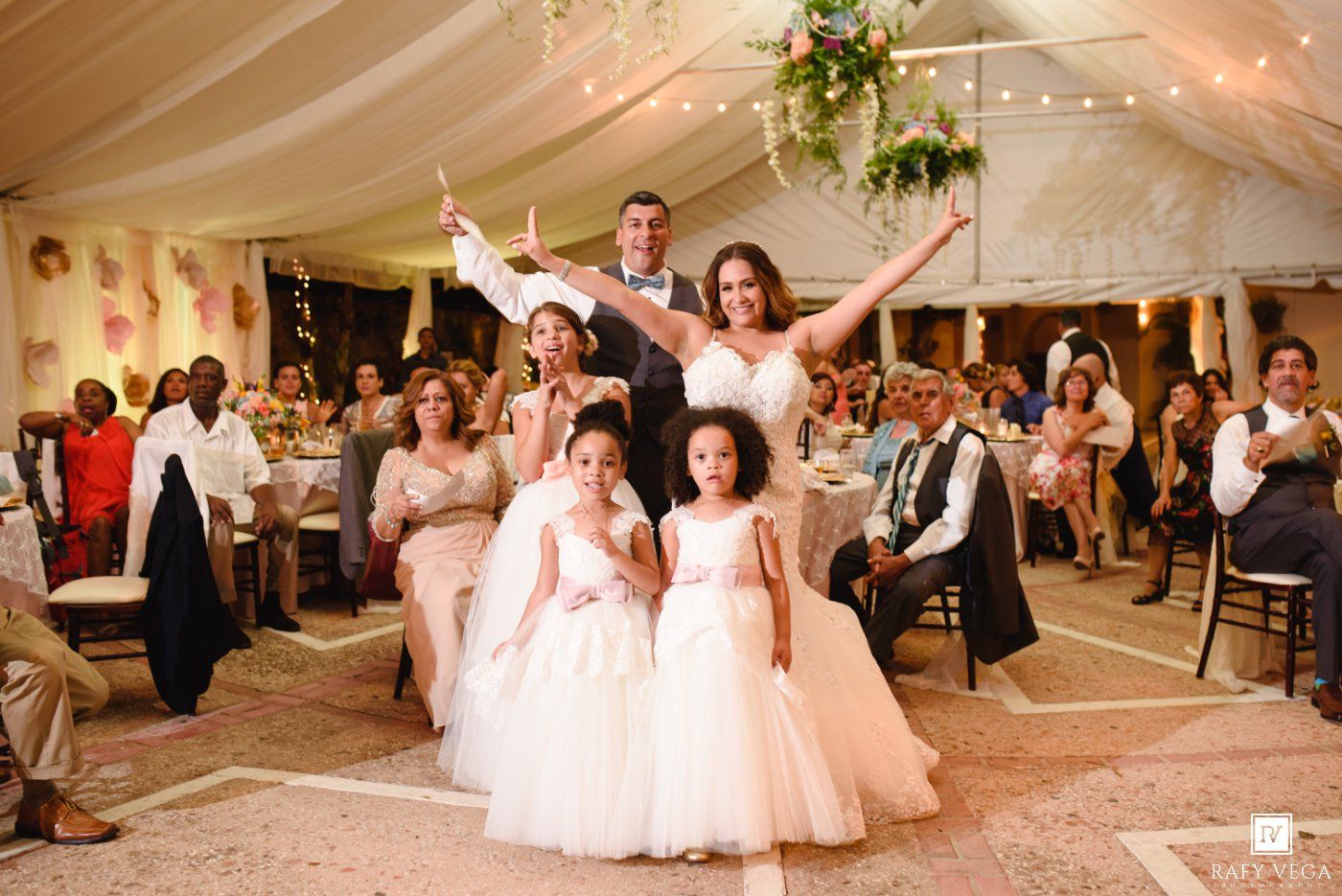 Joyful bride and groom celebrate with flower girls in white dresses at elegant tented wedding reception.