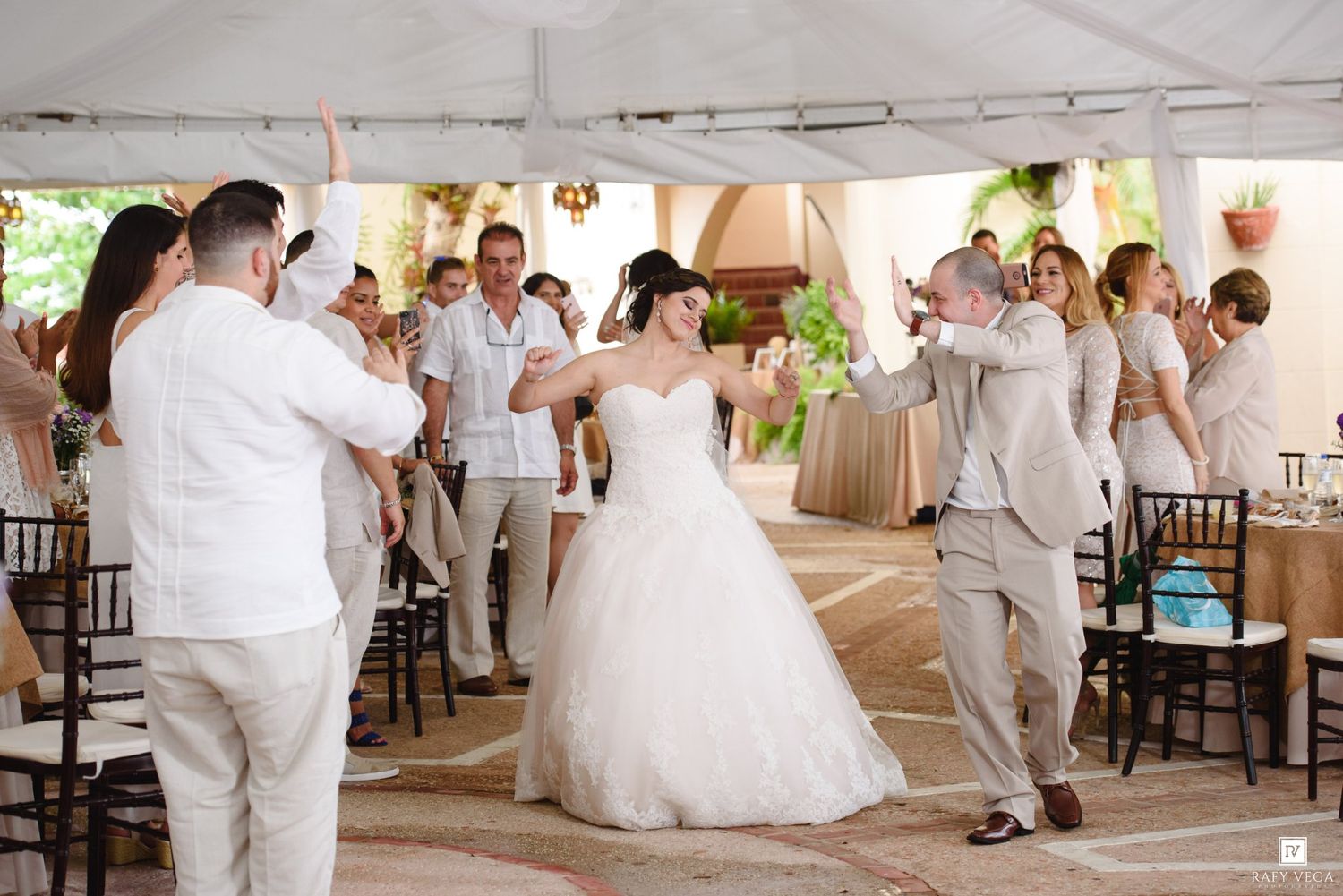 Bride and groom share first dance surrounded by cheering guests at an outdoor wedding reception under a white tent.