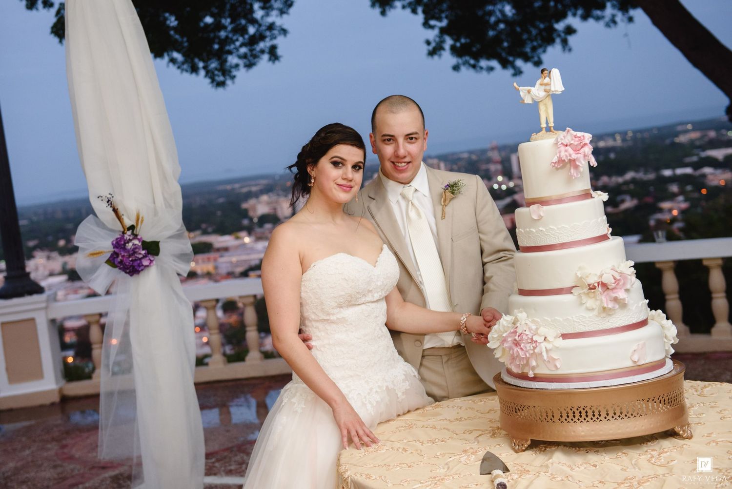 Smiling bride and groom pose beside a tall elegant white wedding cake with pink flowers at dusk reception.