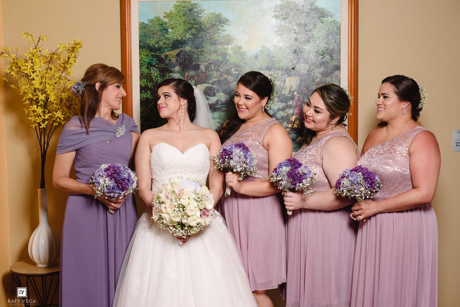 Bride in white gown with bridesmaids in lavender dresses holding purple and white bouquets indoors.