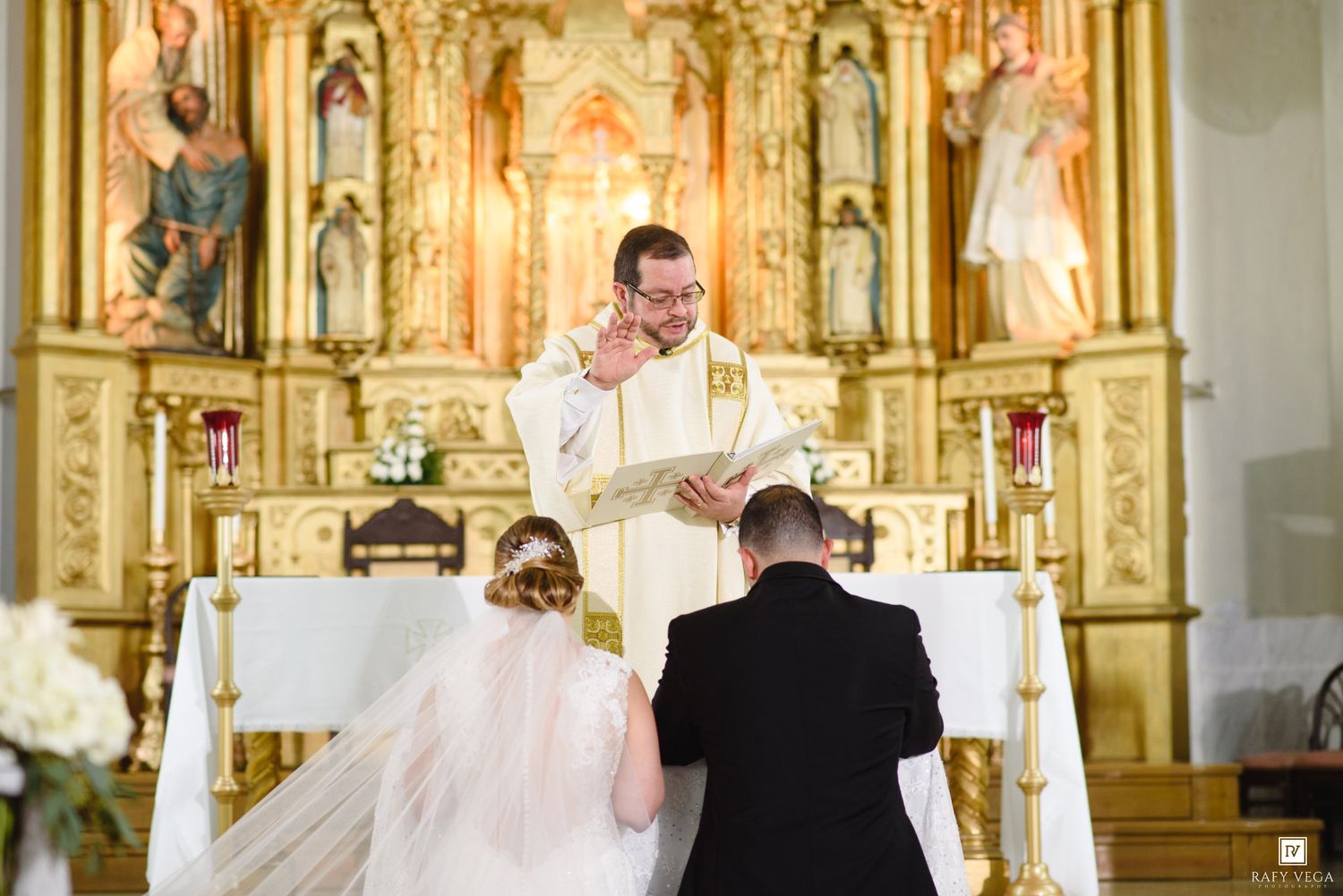 Priest officiating a wedding ceremony at an ornate gold altar in a Catholic church with bride and groom kneeling.