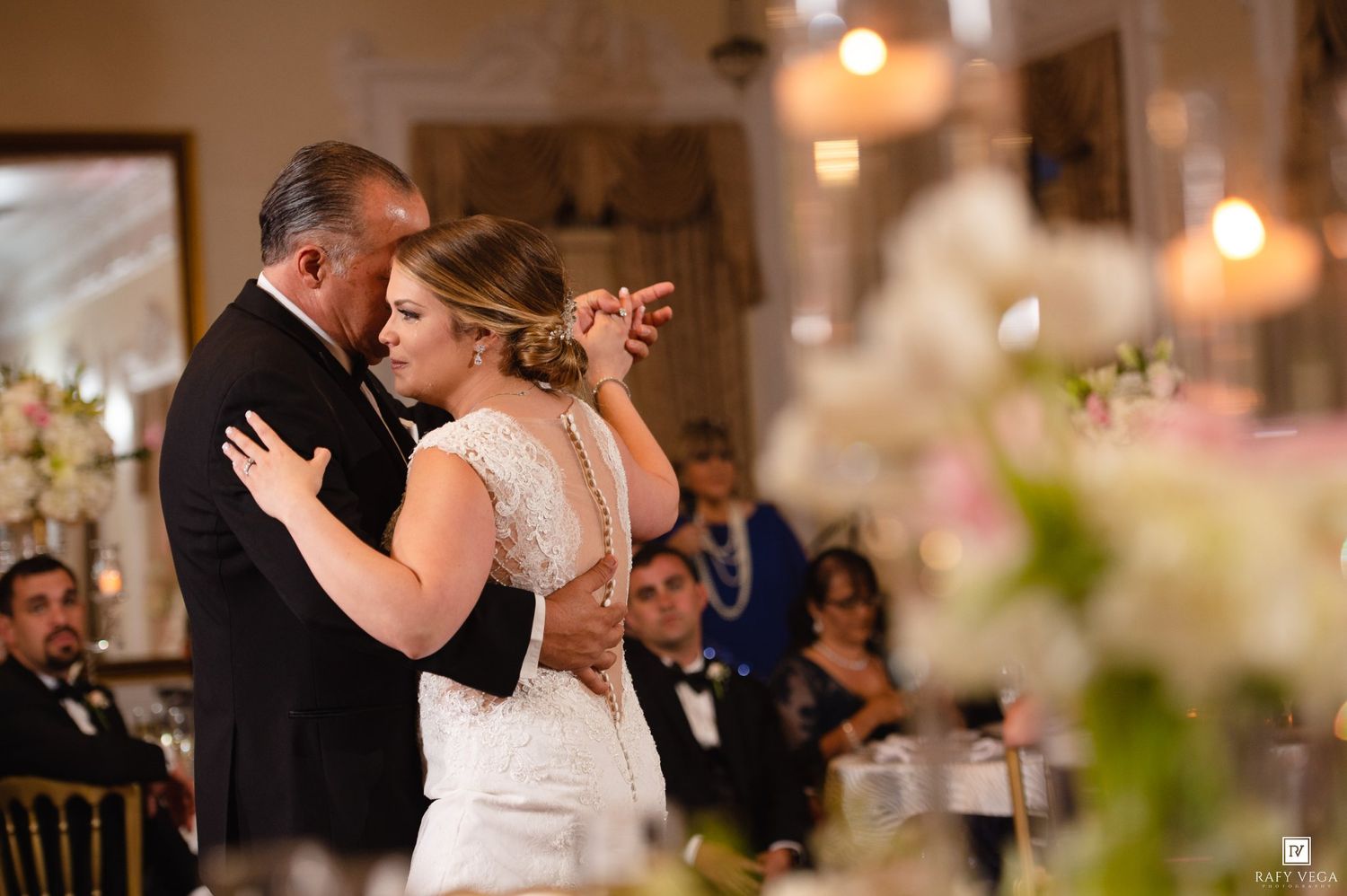 Bride and father share an emotional first dance at elegant wedding reception with floral arrangements and warm lighting.