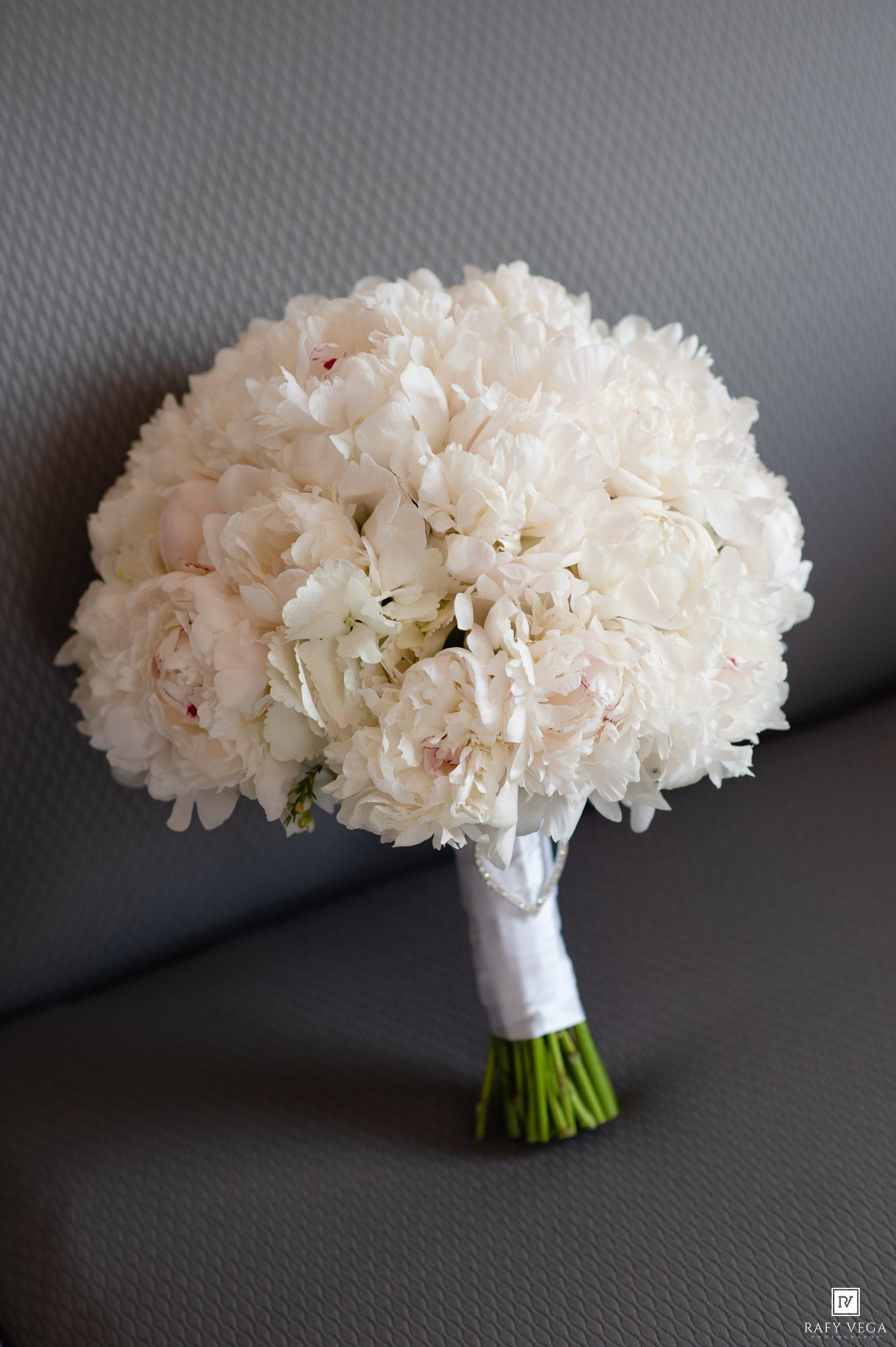 Elegant white peony bridal bouquet with ribbon wrap resting on gray chair for wedding ceremony.