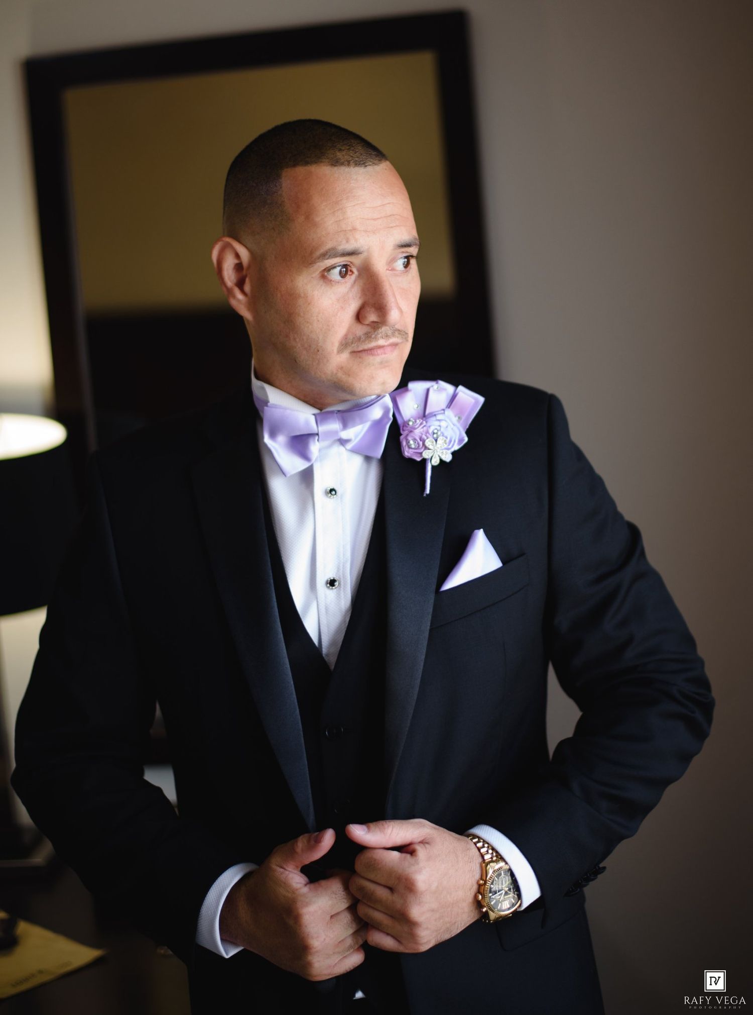 Groom in black tuxedo with lavender bow tie and boutonniere adjusting jacket indoors.