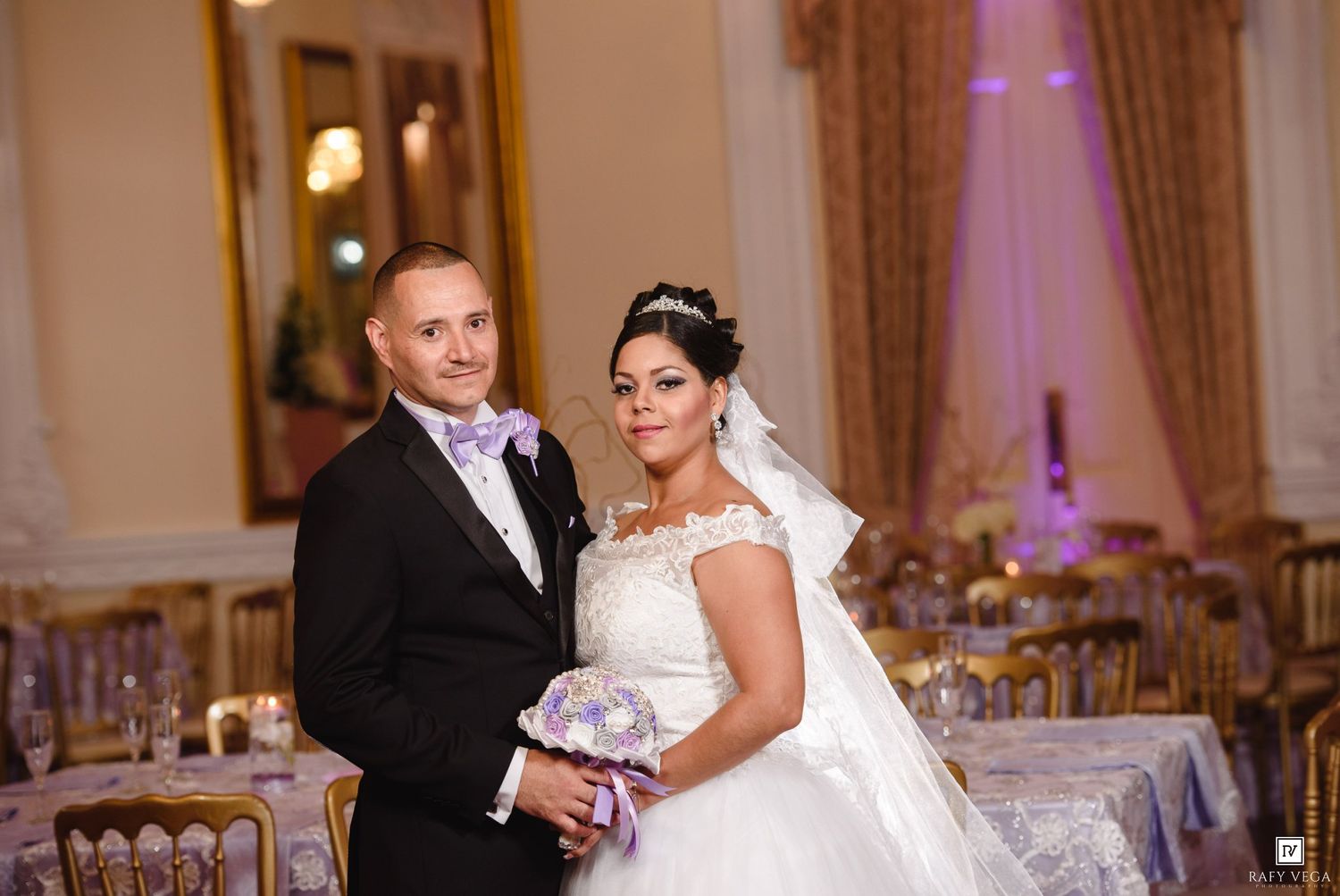Bride in white lace gown and groom in black tuxedo with lavender tie pose together in elegant wedding reception hall.