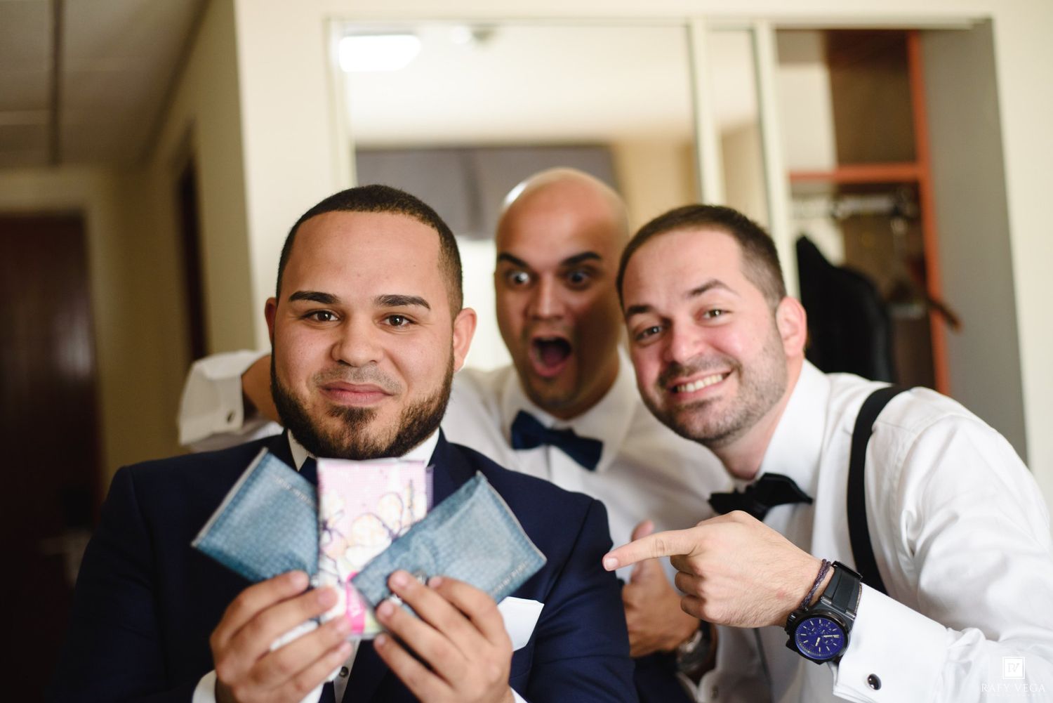 Three groomsmen in bow ties and suspenders pose playfully holding pocket squares before a wedding ceremony.