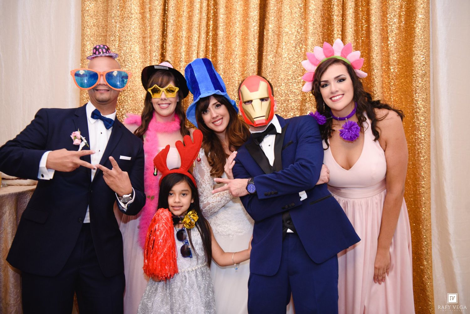 Wedding guests in suits and formal wear pose with colorful masks and photo booth props at elegant gold reception venue.
