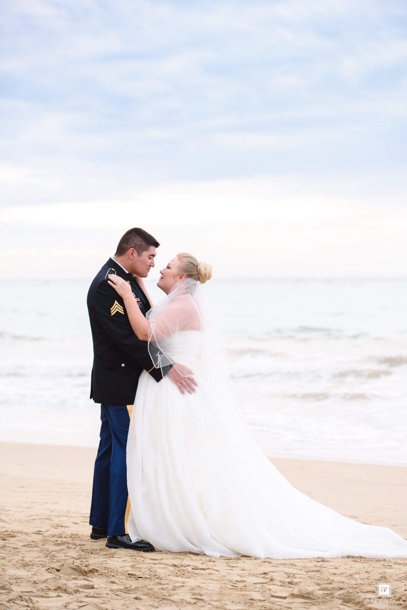 Military groom and bride embrace on sandy beach in wedding attire, ocean waves in background.