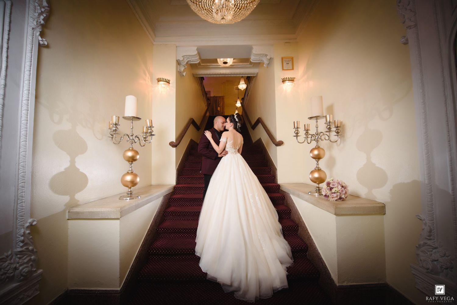 Bride and groom share a romantic embrace on a grand staircase in an elegant wedding venue with ornate decor.