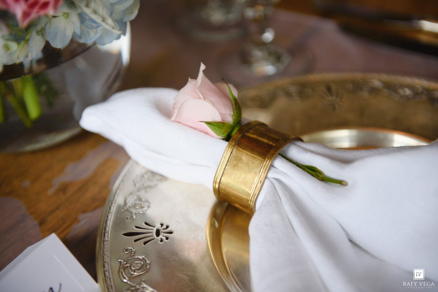 Elegant gold charger plate with white linen napkin, gold ring, and pink rose bud on a wooden wedding reception table.