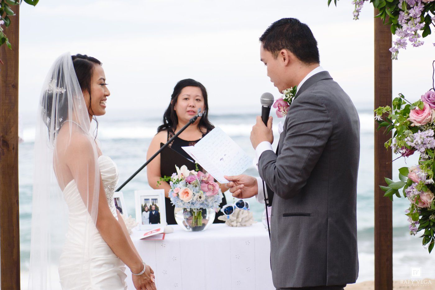 Groom reads vows to bride at beach wedding ceremony with officiant and floral arrangements at altar.
