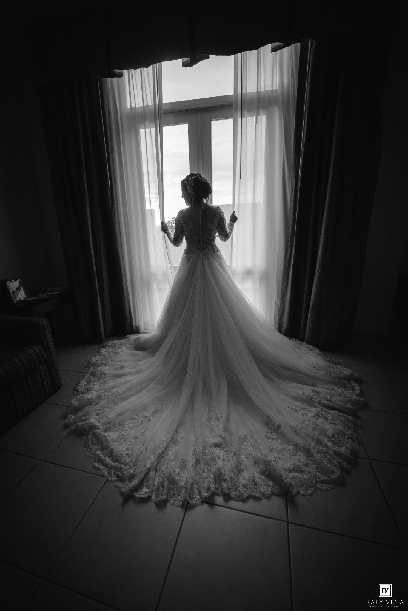 Bride in elegant lace wedding gown with long train standing before window in black and white photo.