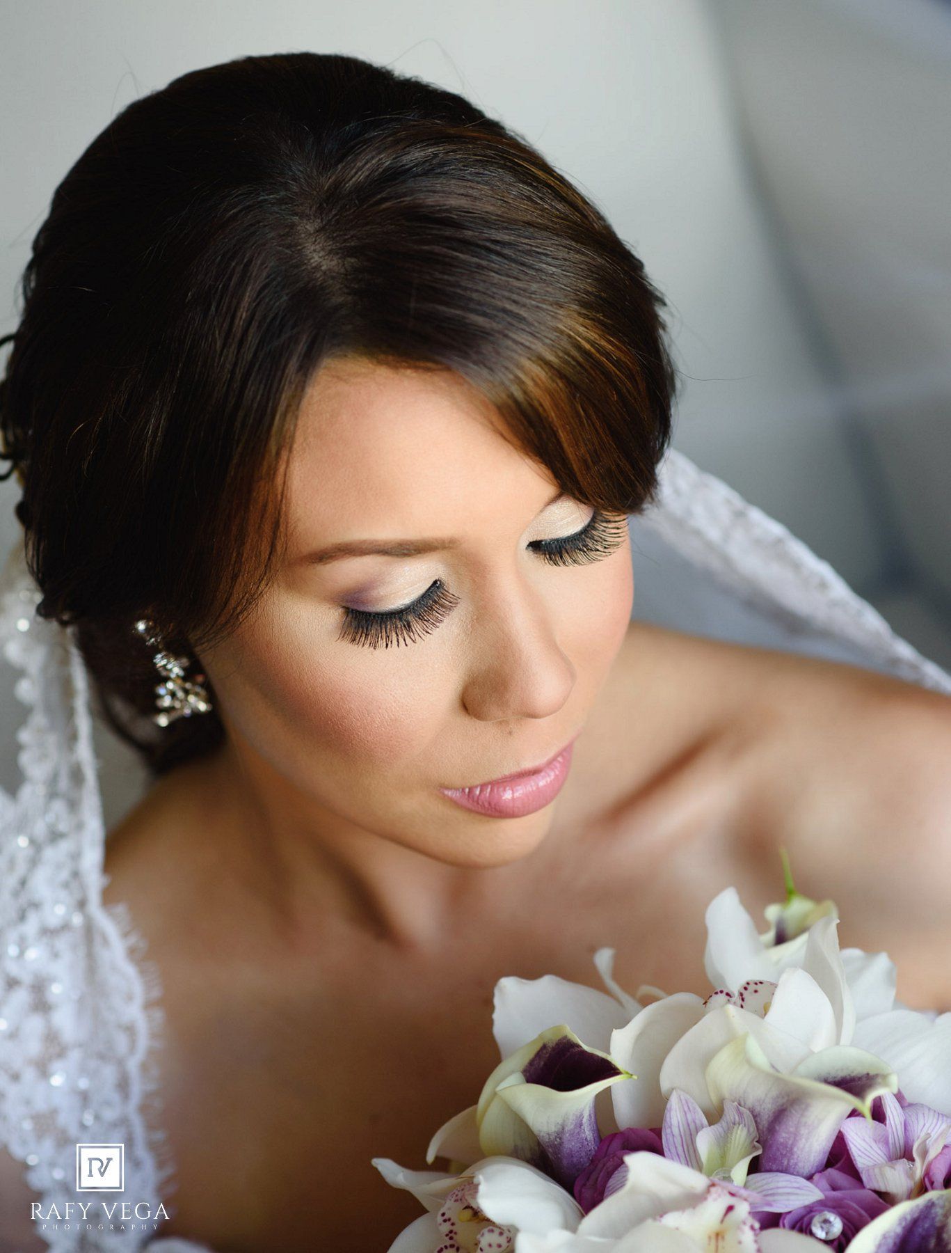 Bride with elegant updo, dramatic lashes, and soft glam makeup holding a white and purple orchid bouquet.