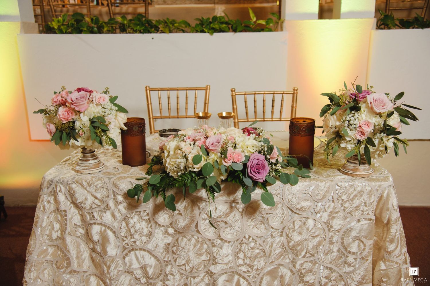 Elegant wedding sweetheart table with gold chairs, lush pink and white floral arrangements, and a champagne rosette tablecloth.