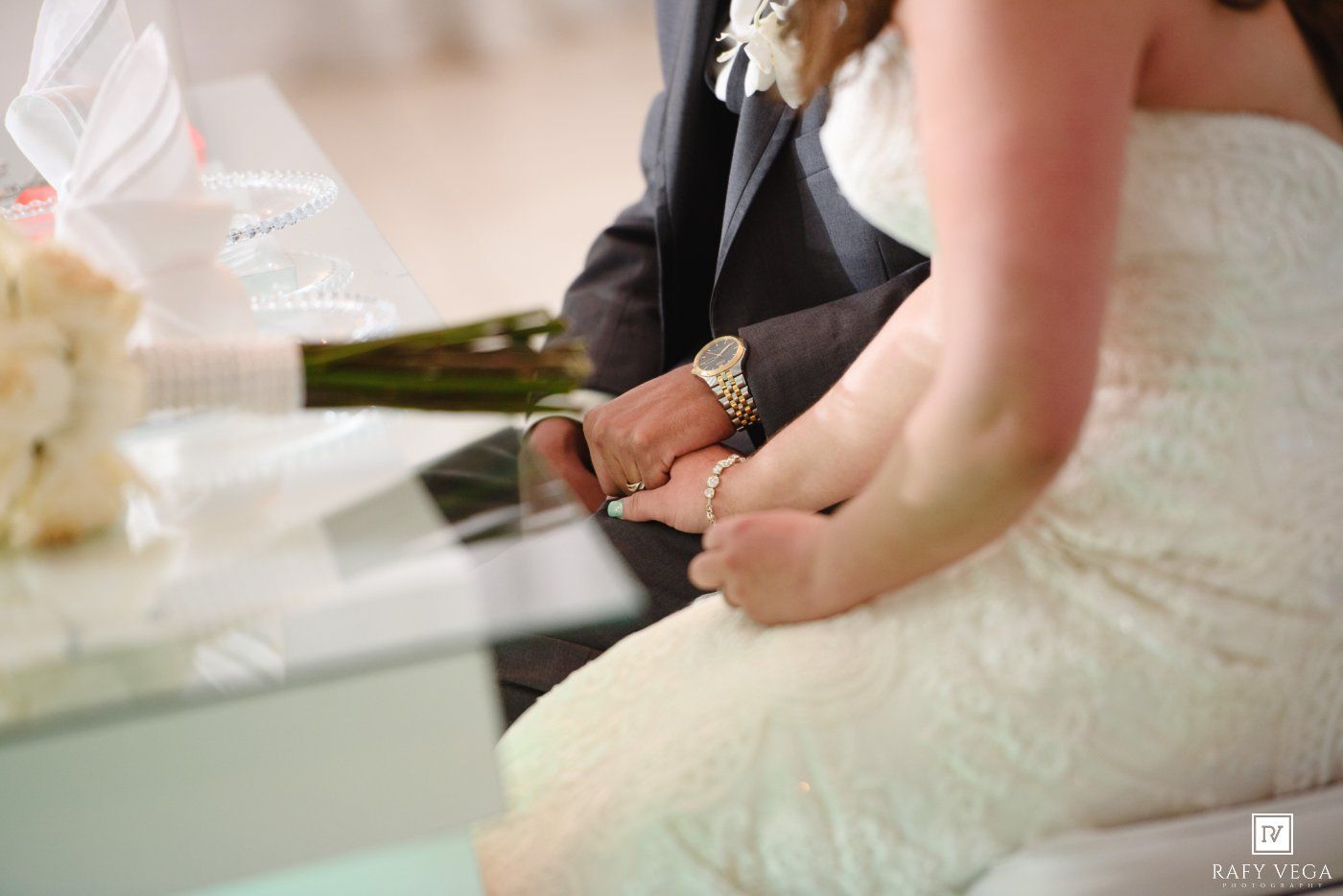 Bride and groom holding hands at wedding ceremony, bride wearing lace gown and groom in dark suit with gold watch.