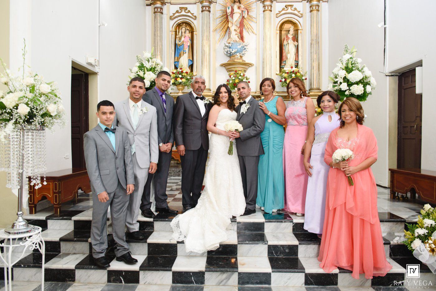 Wedding party poses in a church with bride and groom surrounded by bridesmaids and groomsmen near ornate altar.
