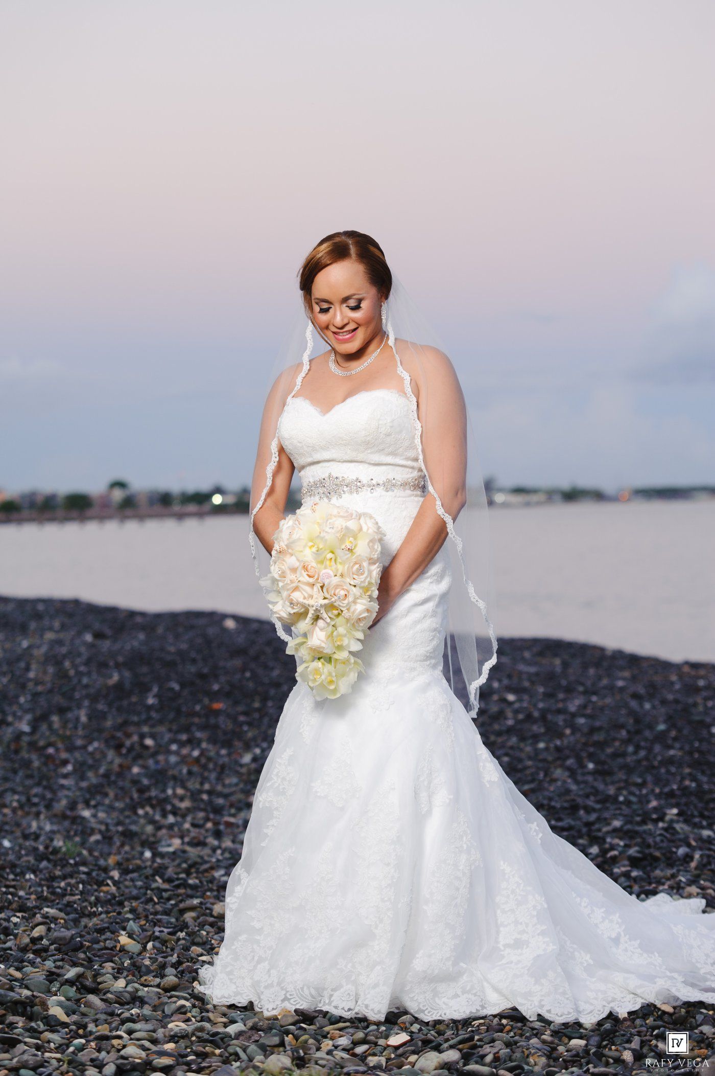 Bride in white mermaid gown holding cascading bouquet on rocky beach at sunset.