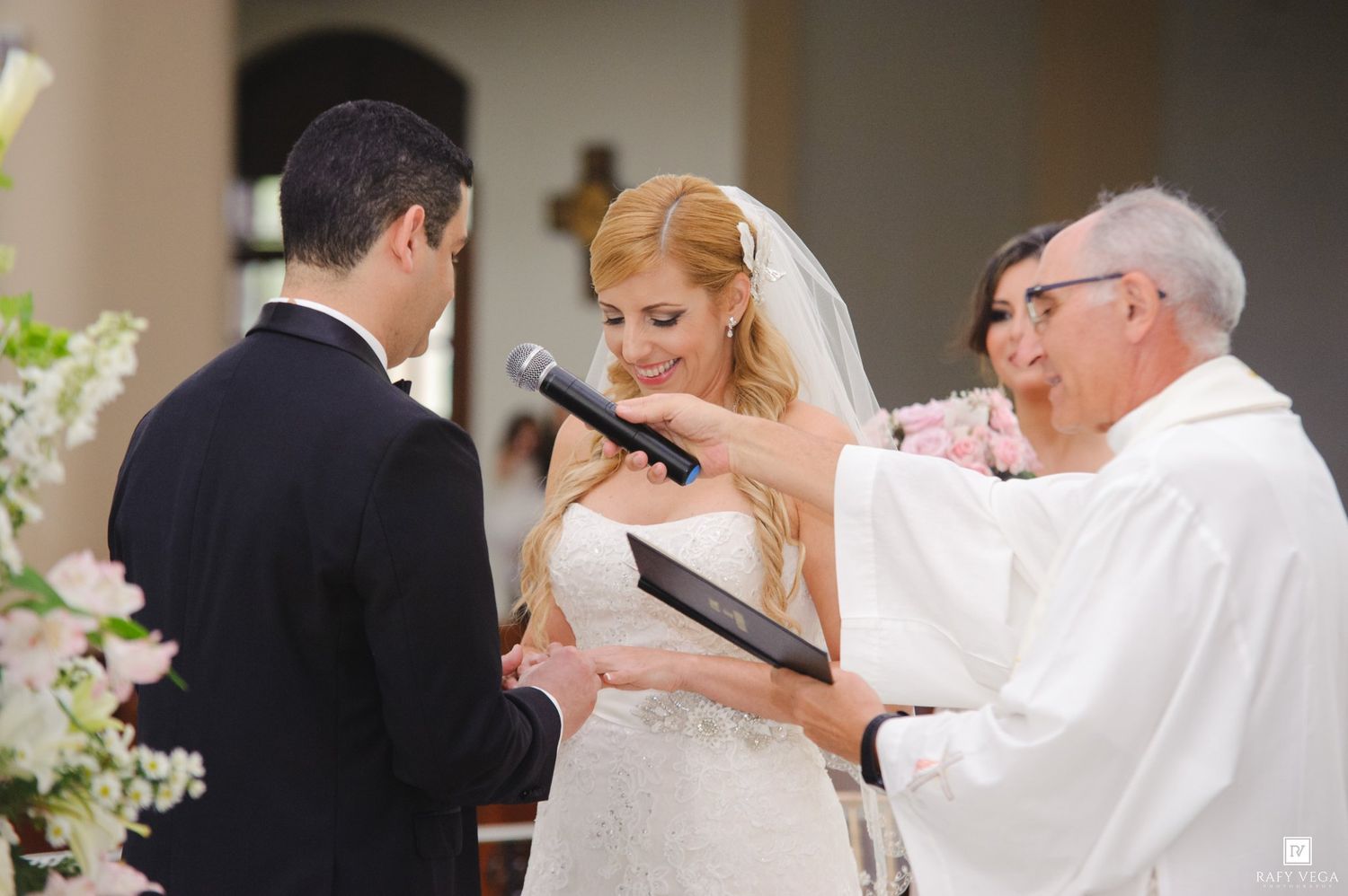 Bride and groom exchange vows at church altar with officiant holding microphone during elegant wedding ceremony.