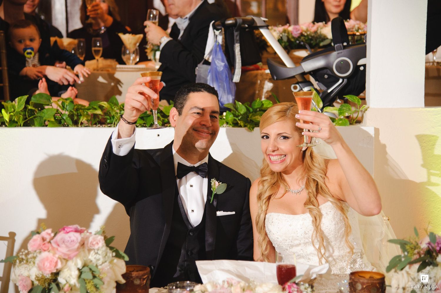 Bride and groom raise champagne glasses in toast at wedding reception with floral centerpieces.