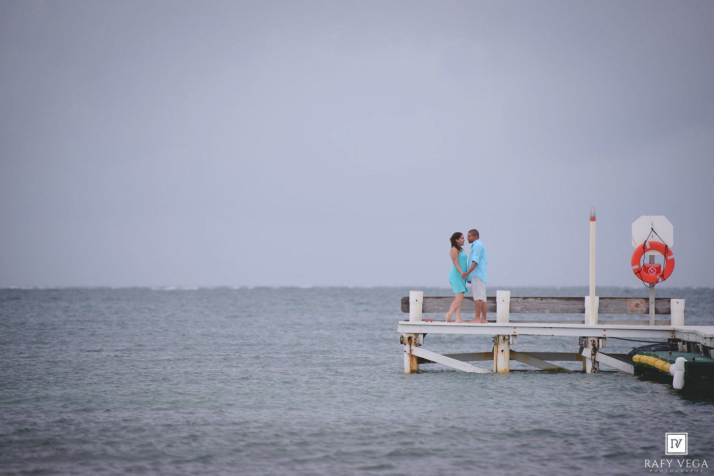 Romantic couple embracing on wooden pier over calm ocean waters under overcast sky.