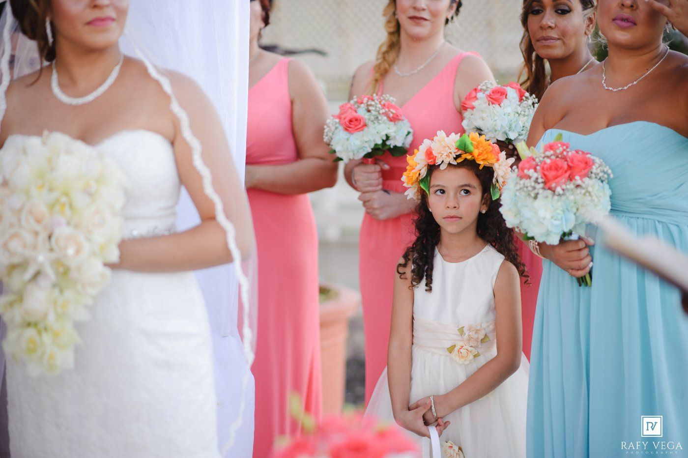 A flower girl in white dress stands with bridesmaids in coral and mint gowns holding colorful bouquets at an outdoor wedding.