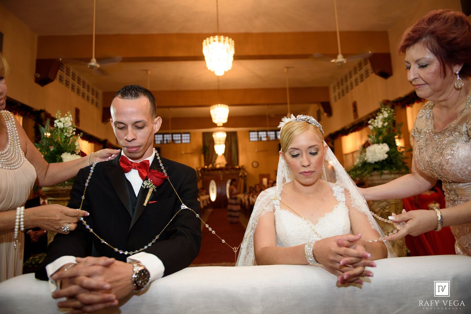 Bride and groom kneeling at church altar during wedding ceremony with floral decorations and guests in background.