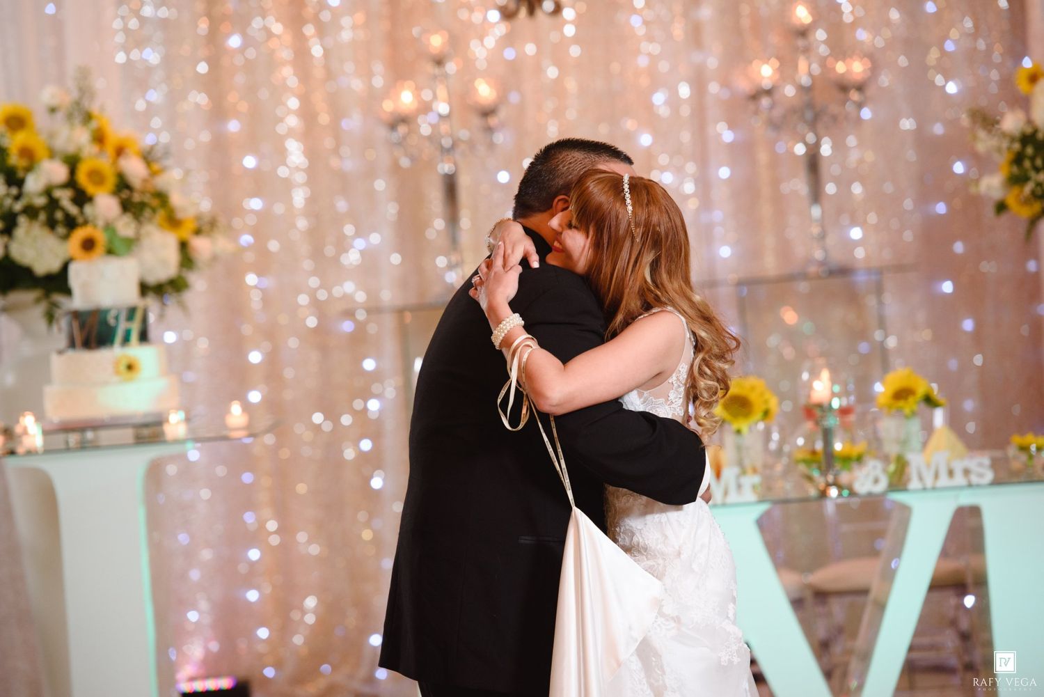 Bride and groom share an emotional first dance embrace at a romantic wedding reception with twinkling lights and sunflowers.