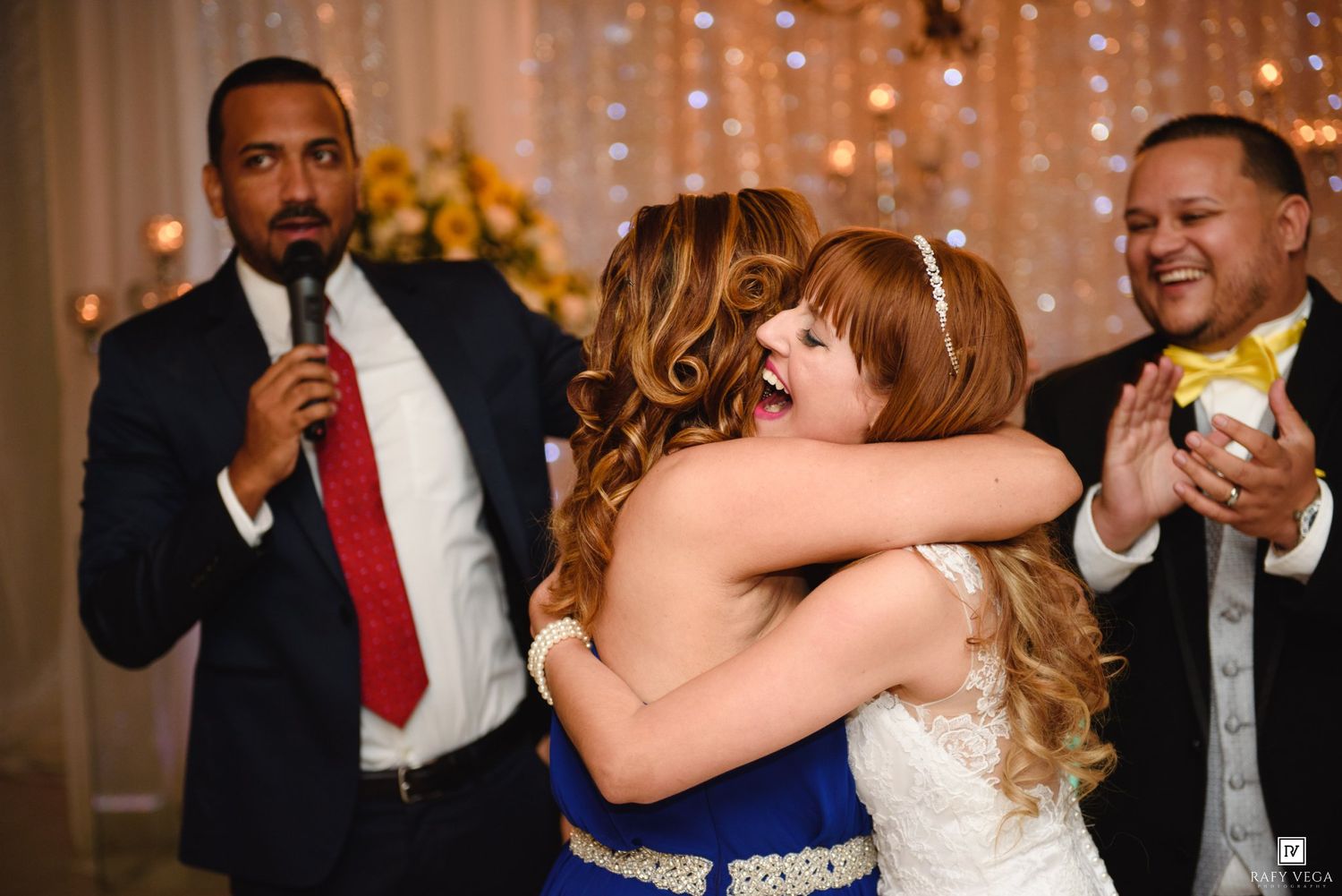 Two women share a warm hug at a wedding reception while guests celebrate in the background.