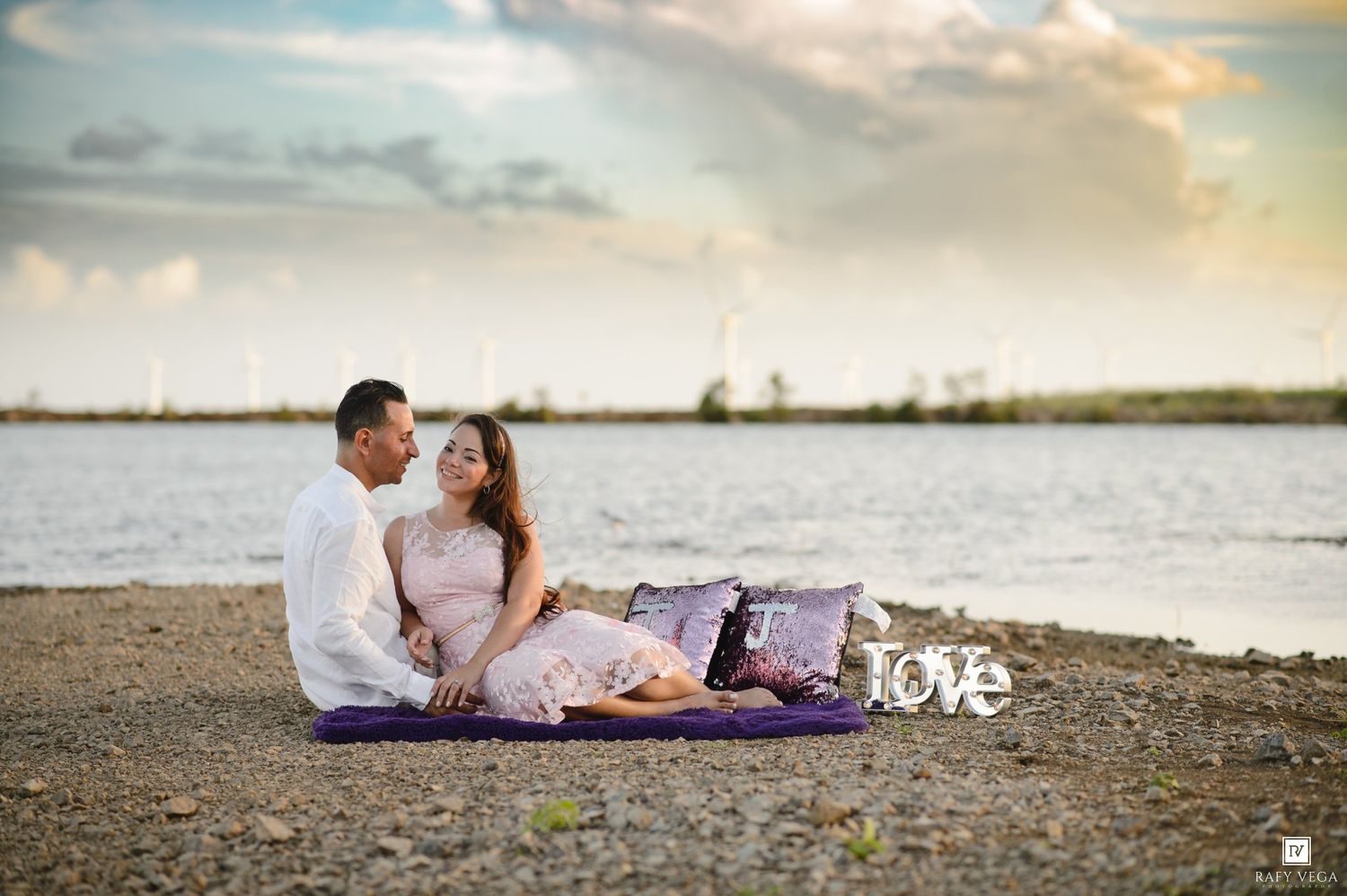 Couple sitting on purple blanket by waterfront with love sign, romantic beach engagement photo at sunset.