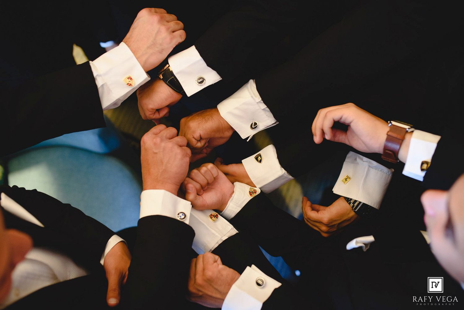 Groomsmen in black tuxedos with white cuffs and gold cufflinks doing a group fist bump at a wedding.