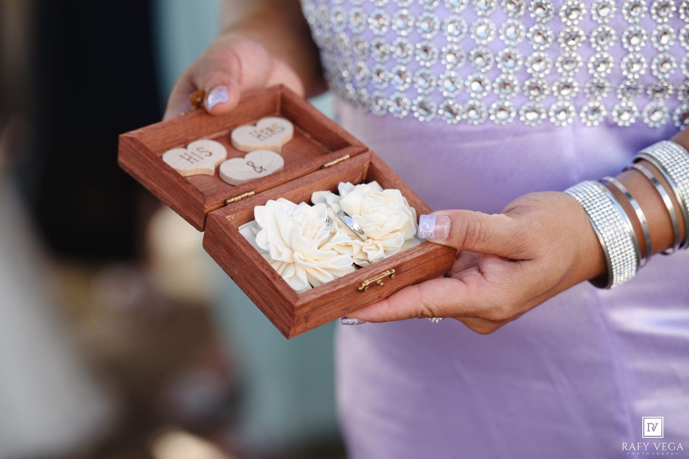 Woman holding a wooden ring box with white flowers and wedding rings at an outdoor ceremony.
