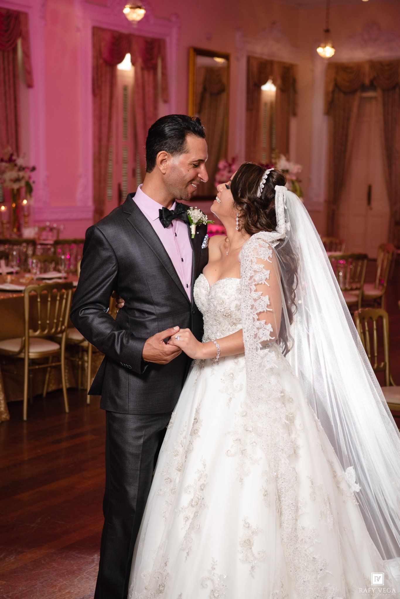 Bride and groom share a romantic first dance in an elegant pink ballroom, bride wearing a lace ball gown and veil.