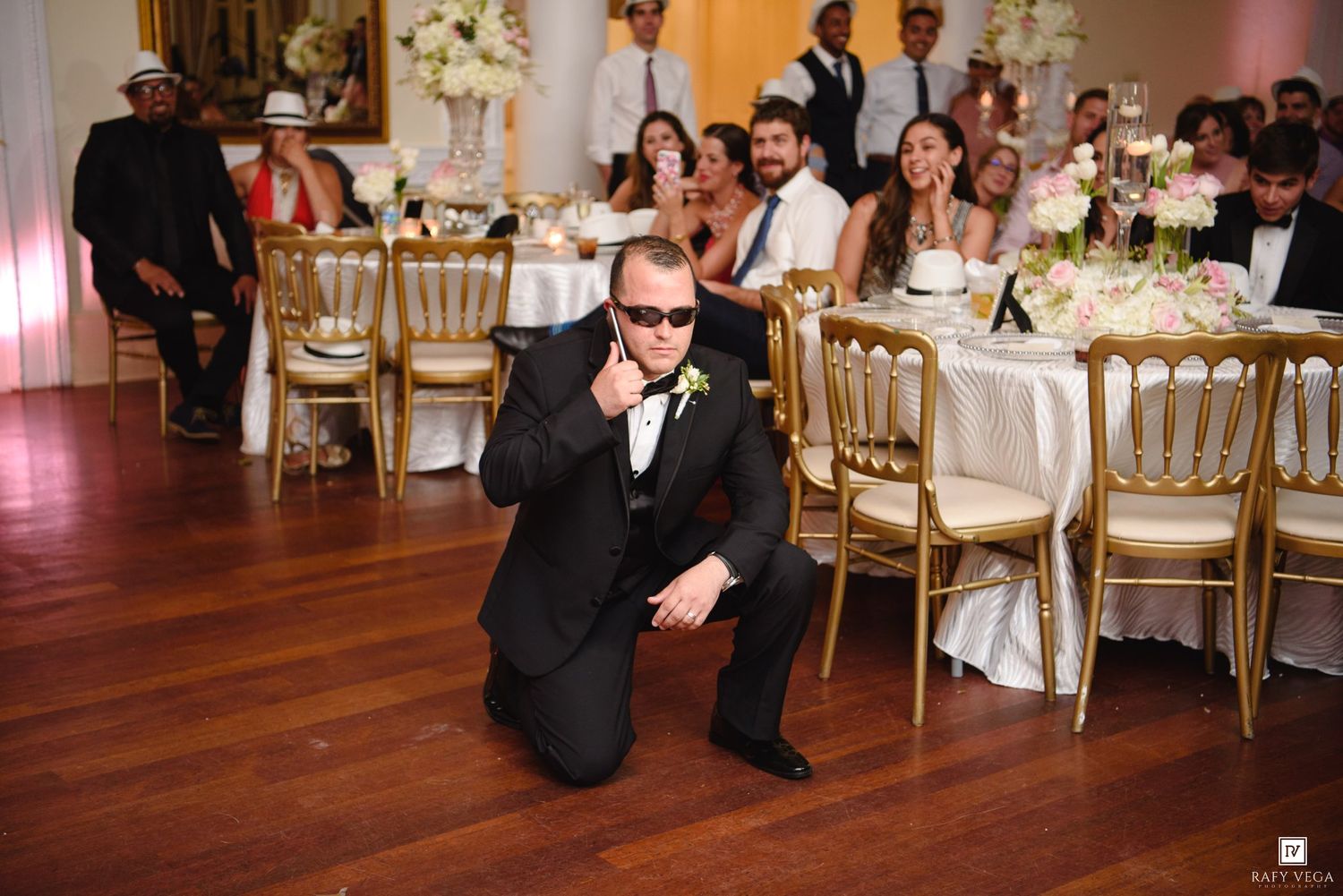 A man in a black tuxedo kneels on a dance floor at an elegant wedding reception with gold chairs and guests.