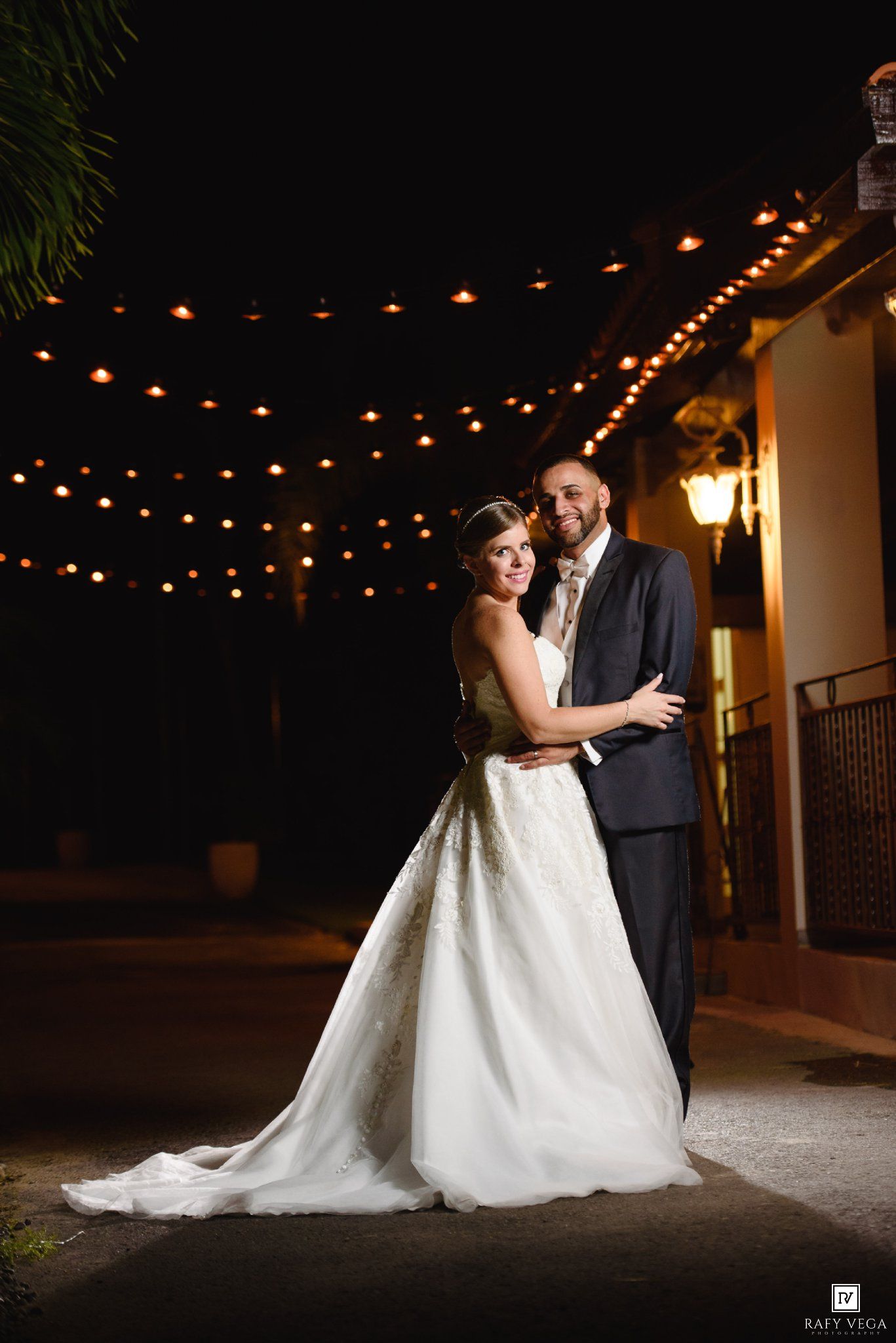 Bride and groom embrace under string lights at night during outdoor wedding reception.
