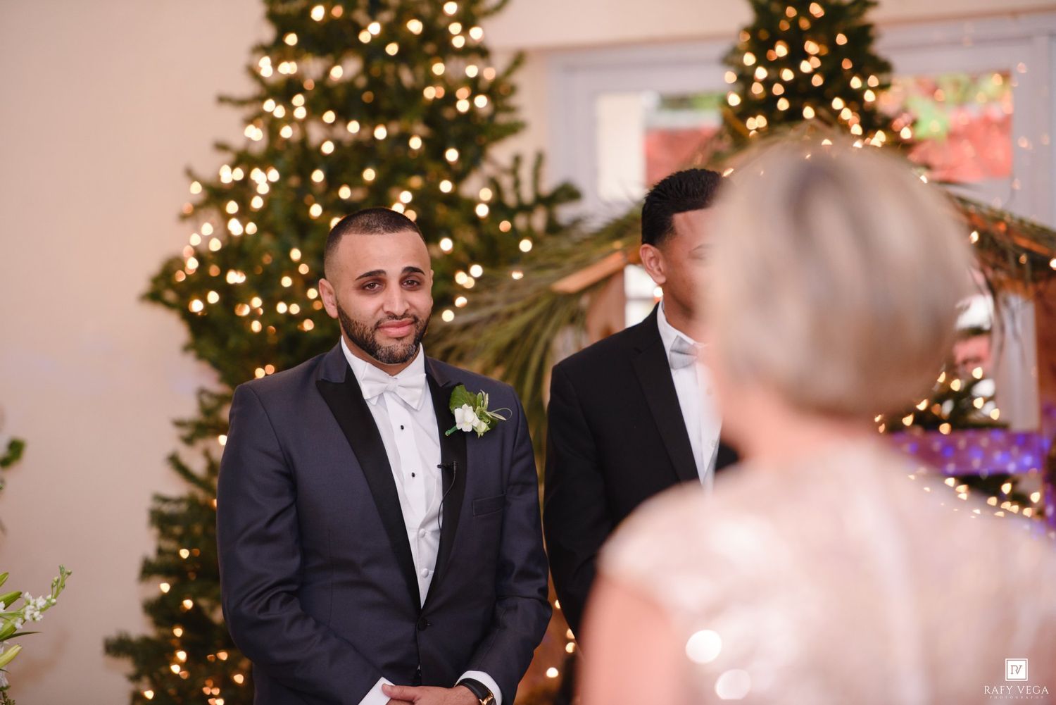 Groom smiling at bride during Christmas wedding ceremony with decorated tree and warm lights in background.