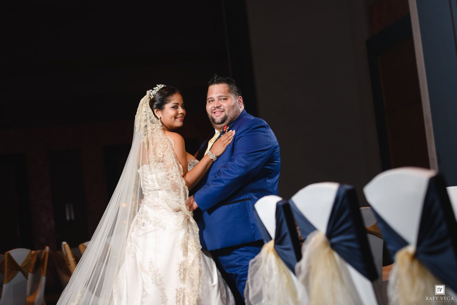 Bride in white gown and veil shares first dance with groom in navy blue suit at elegant wedding reception.